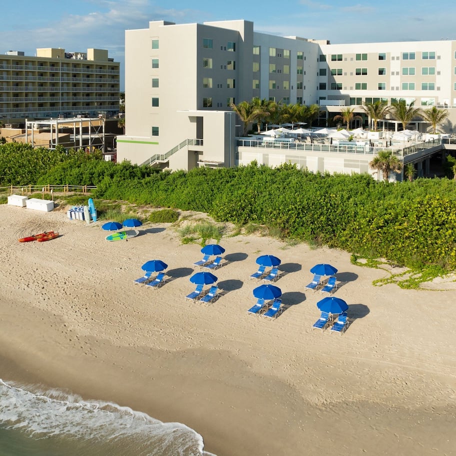 A beach with rows of blue umbrellas and lounge chairs, a hotel building in the background, and a clear view of the ocean.