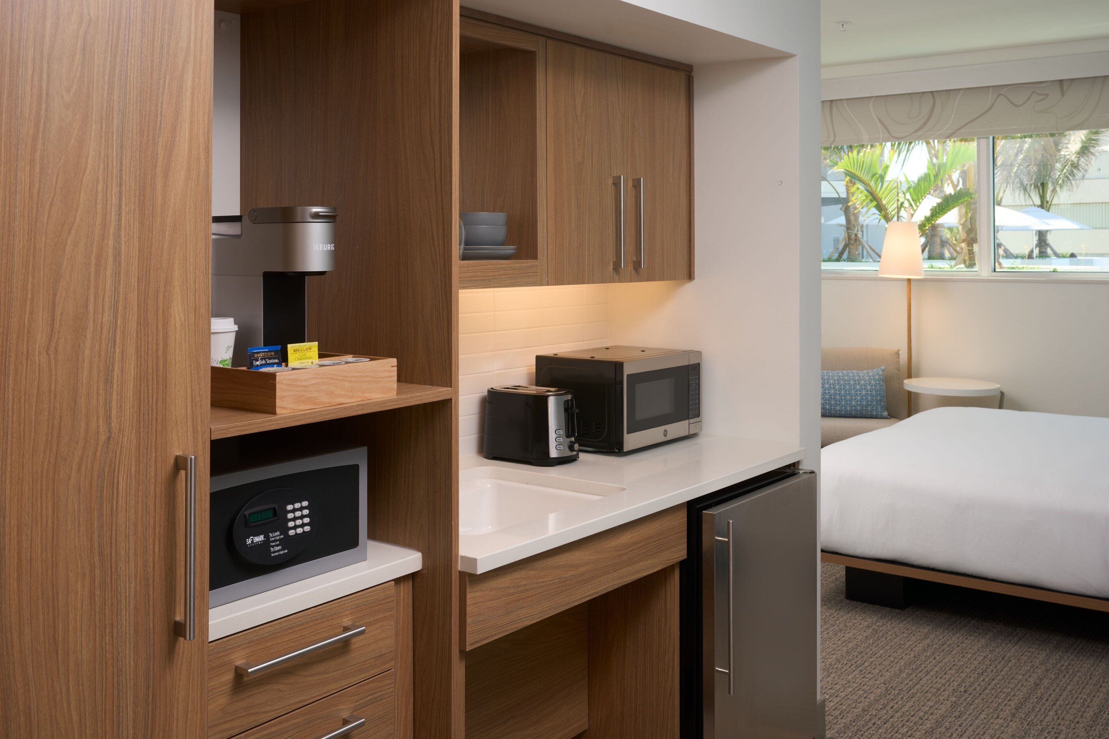 Une chambre d’hôtel compacte comprenant un coin cuisine, un lit et une fenêtre donnant sur des palmiers.