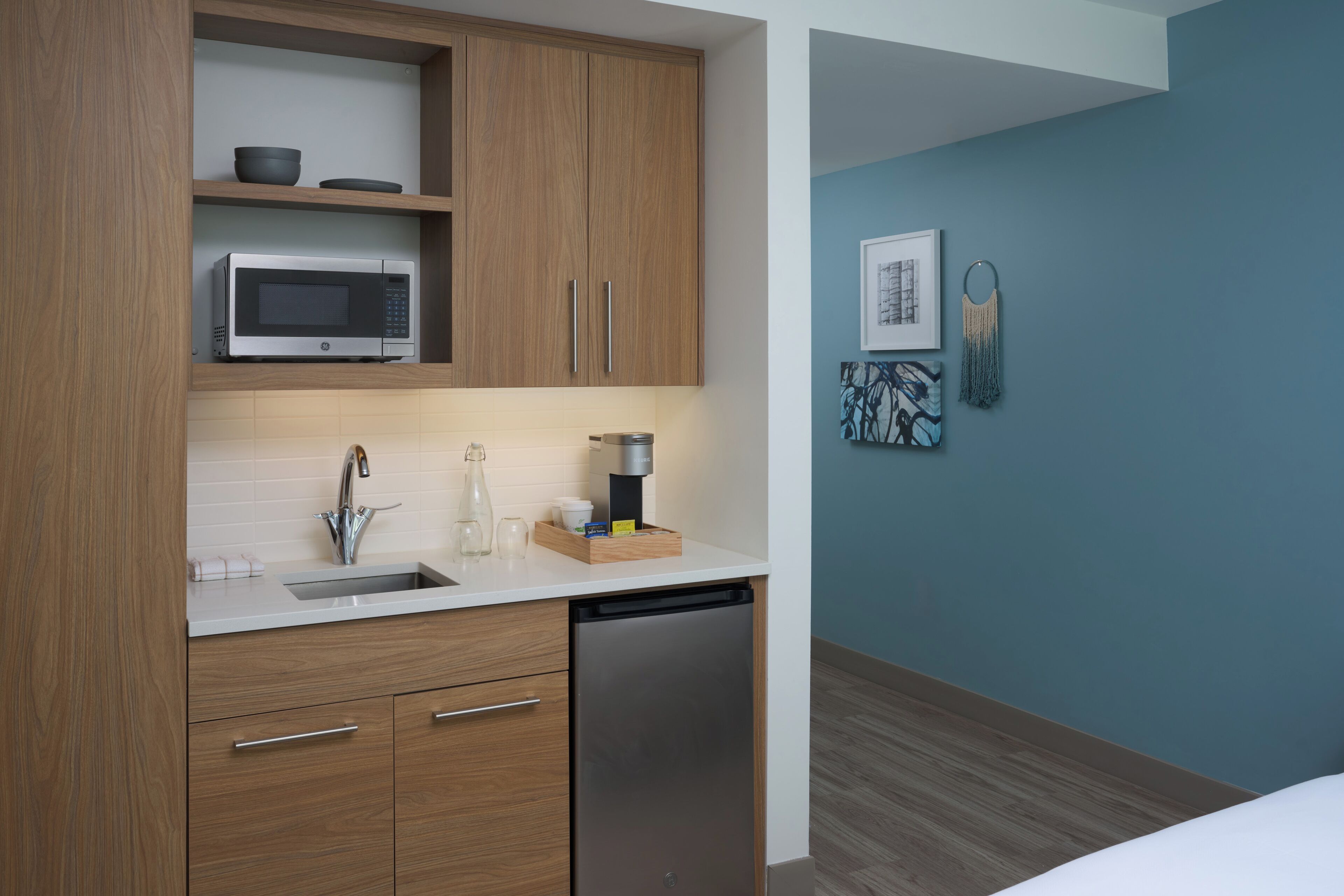 A modern kitchen with wooden cabinets, a microwave, a sink, and a dishwasher. There is a blue wall with artwork and a wooden floor.
