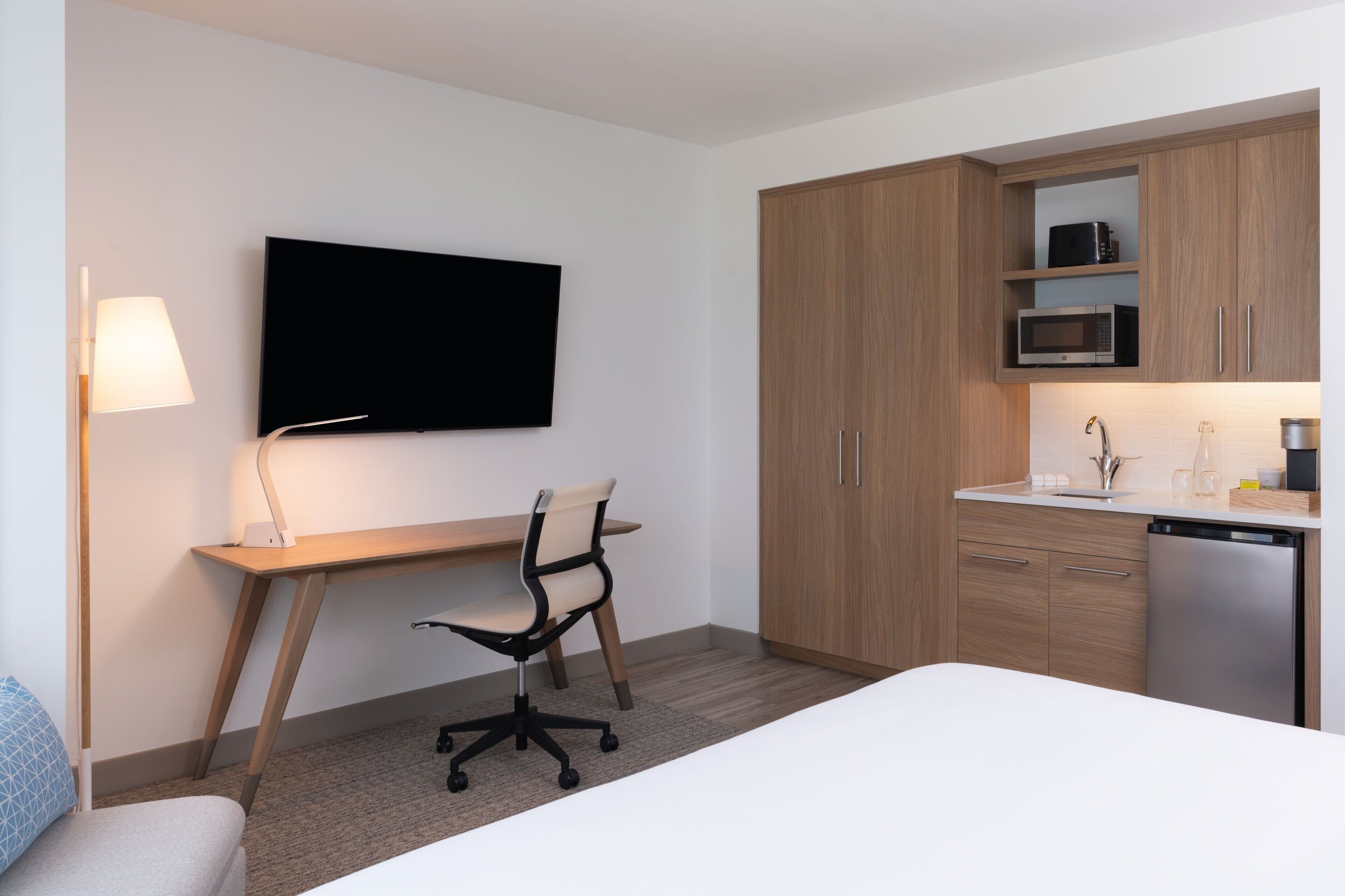 A modern hotel room with a bed, a desk with a chair, a flat-screen TV, wooden cabinets, a microwave, and a dishwasher.