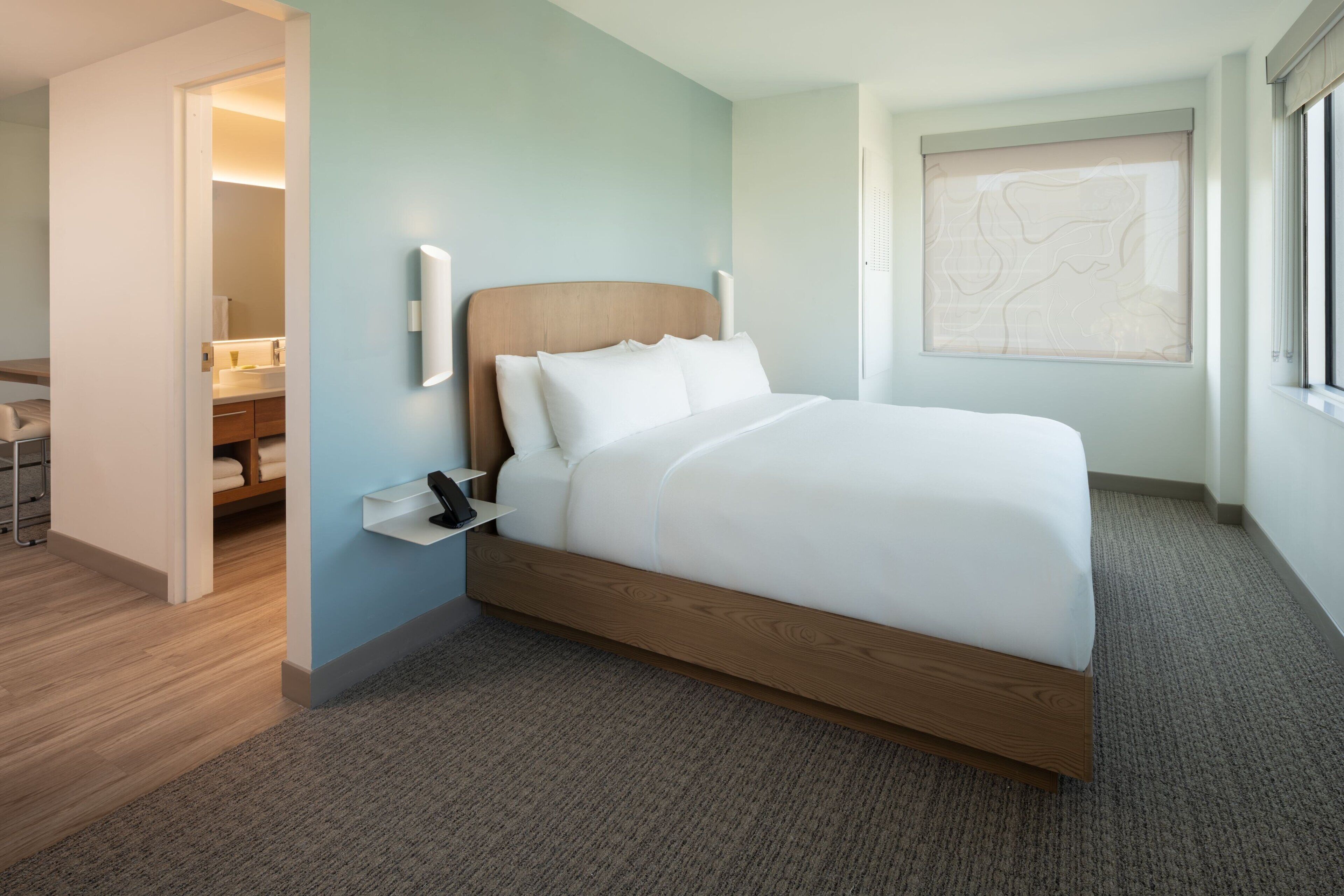 A modern hotel room with a wooden bed, a bedside table, and a large window with a view.