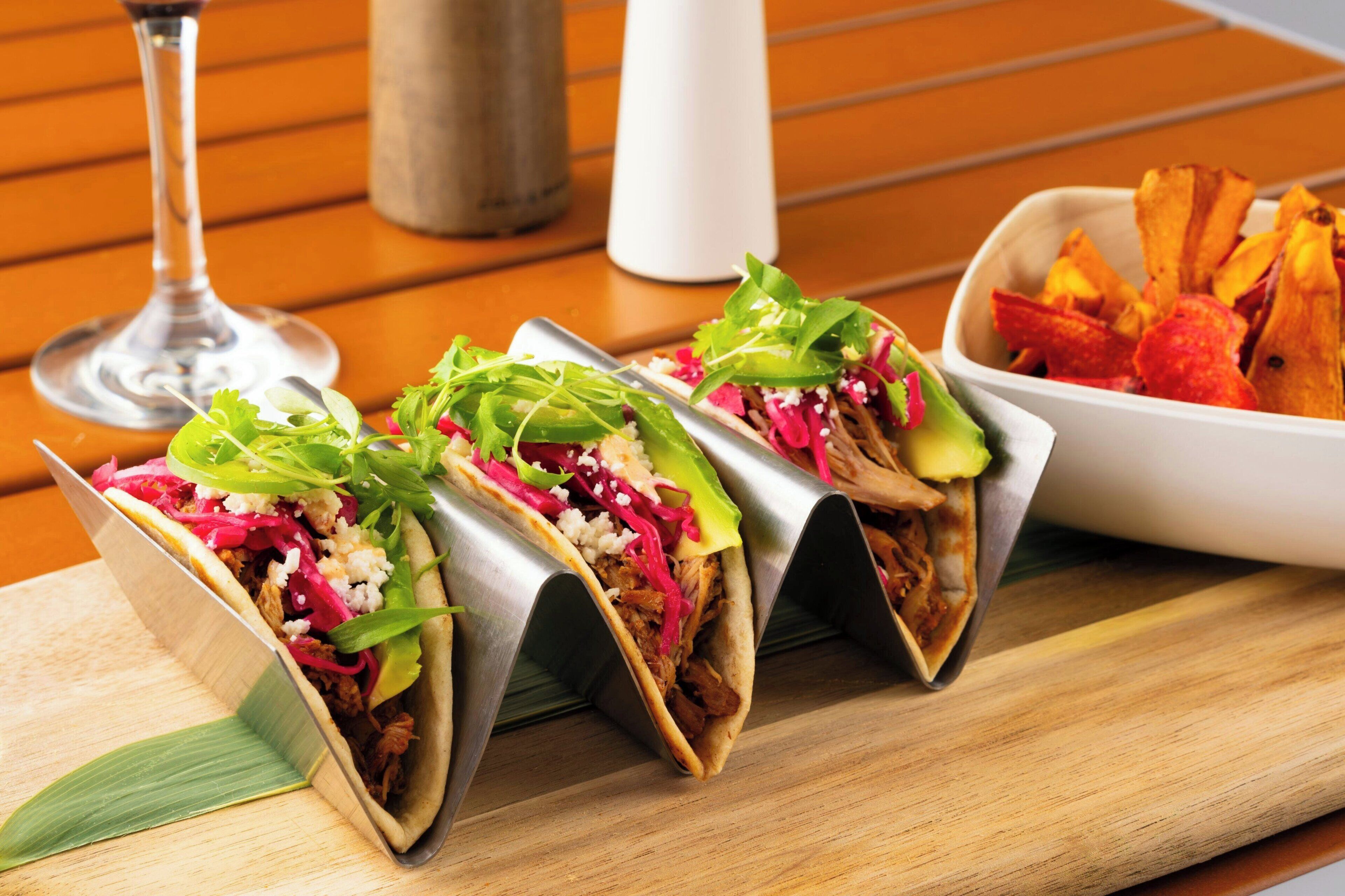 Three tacos on a wooden table with a glass of wine and a bowl of fries.