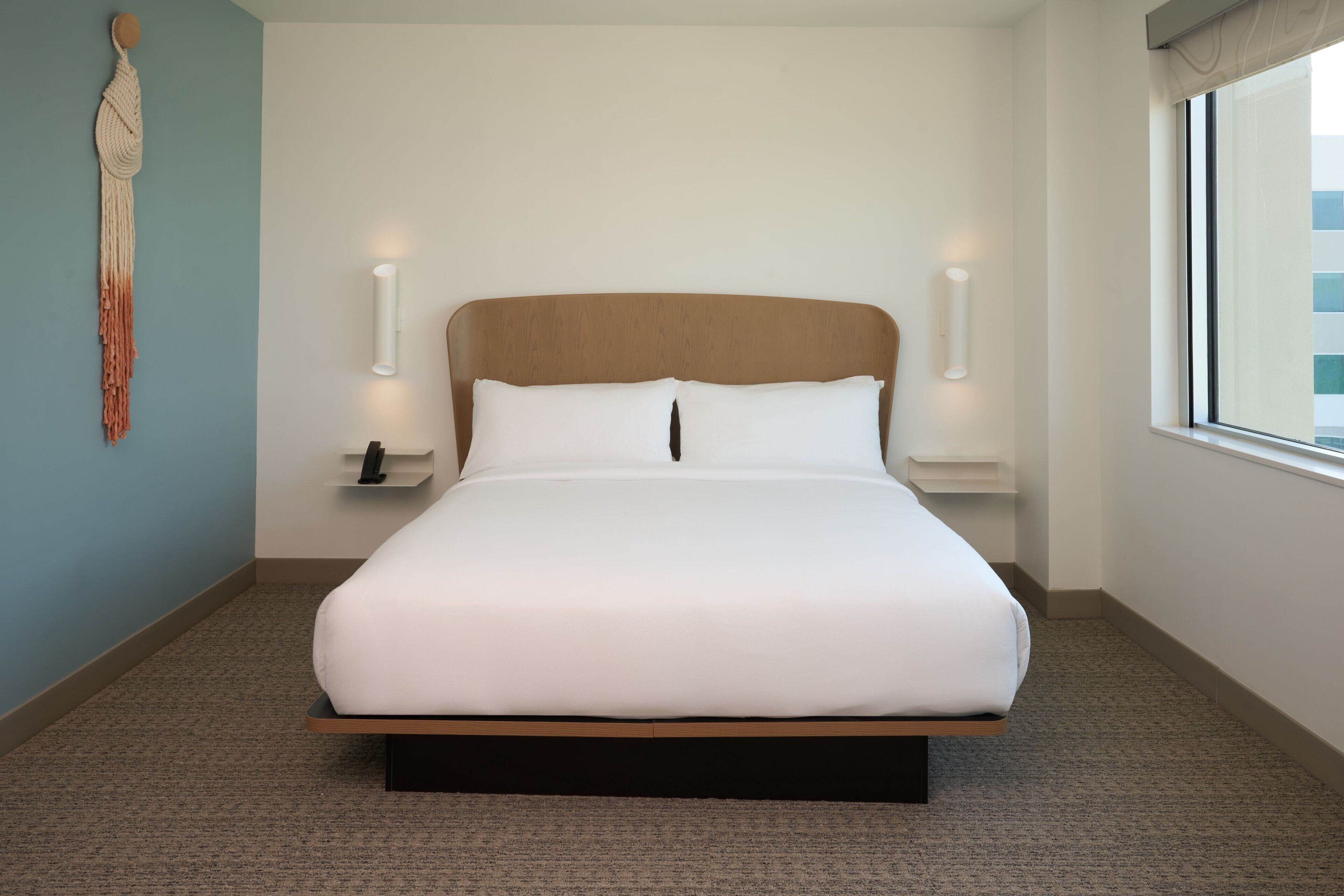 Una cama bien tendida en una habitación de hotel, con un cabecero de madera, dos mesitas de noche con lámparas y un tapiz de macramé en la pared.