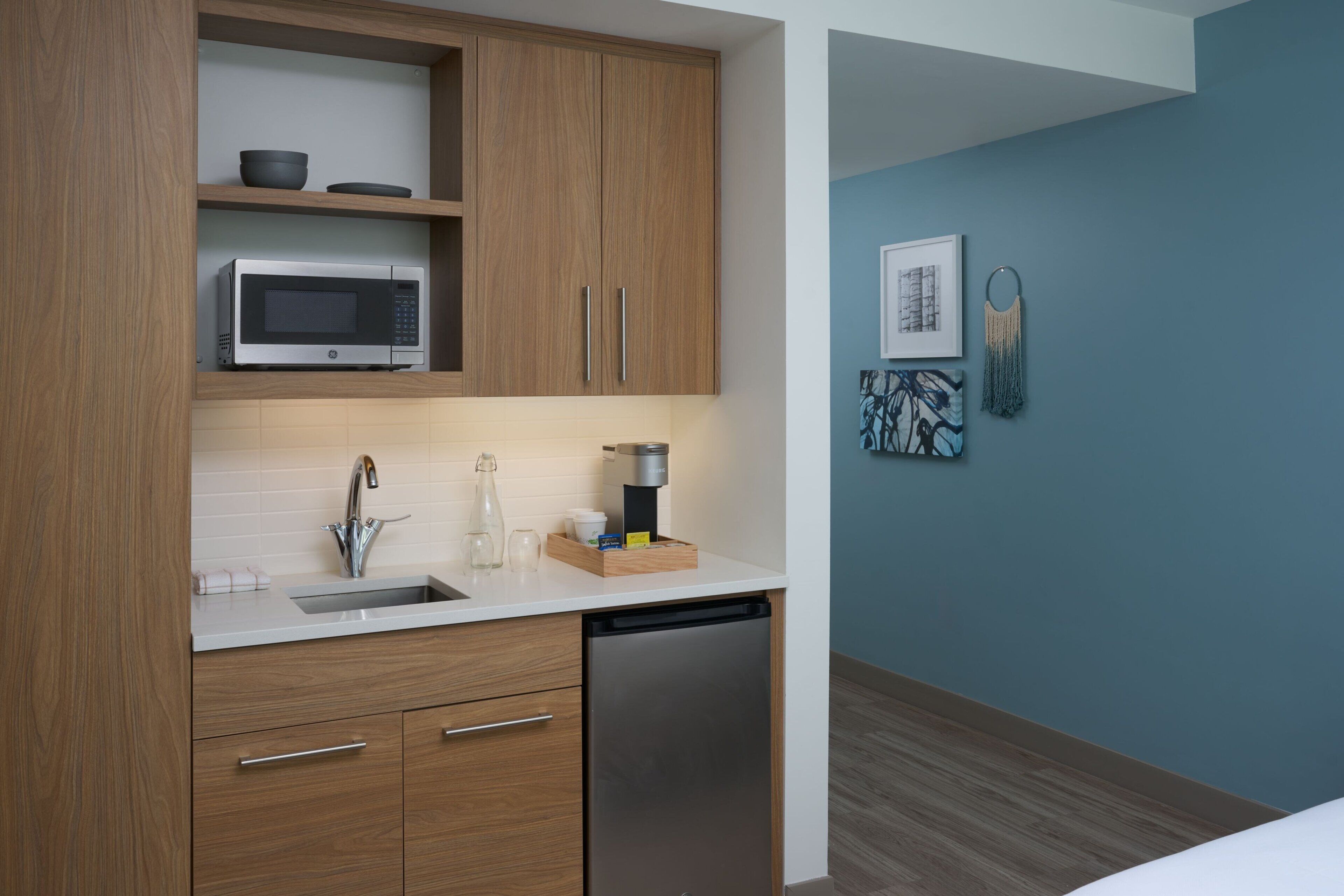 A modern kitchen with wooden cabinets, a microwave, a sink, and a dishwasher. There are decorative items on the counter and artwork on the wall.