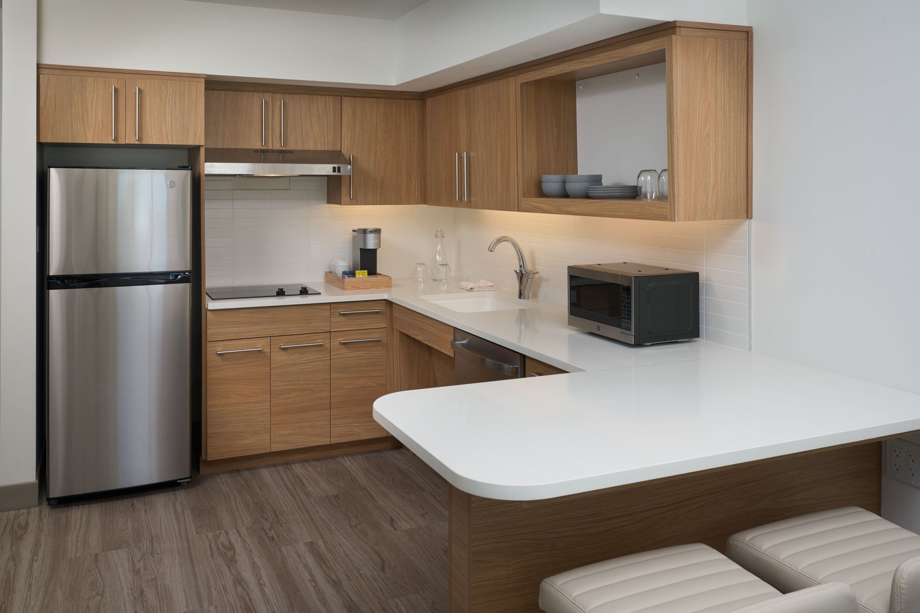 A modern kitchen with wooden cabinets, a stainless steel refrigerator, a microwave, and a white countertop.