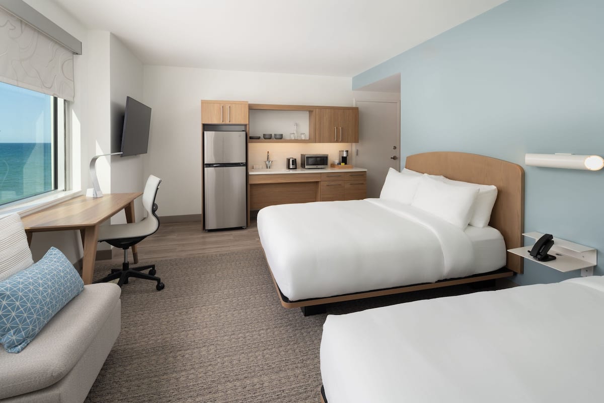 A hotel room with a bed, a desk, a chair, a kitchenette, and a TV.