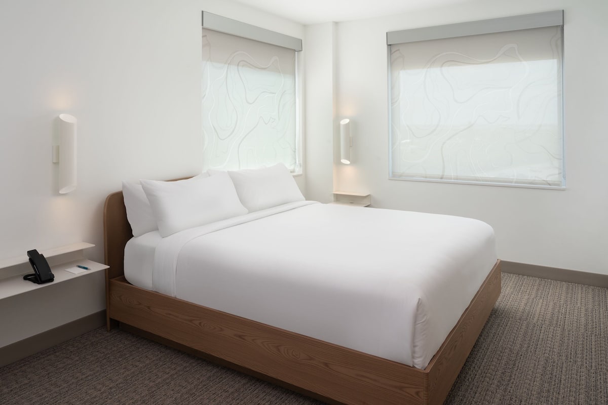 A modern bedroom with a wooden bed, white linens, a bedside table with a phone, and wall-mounted lights.