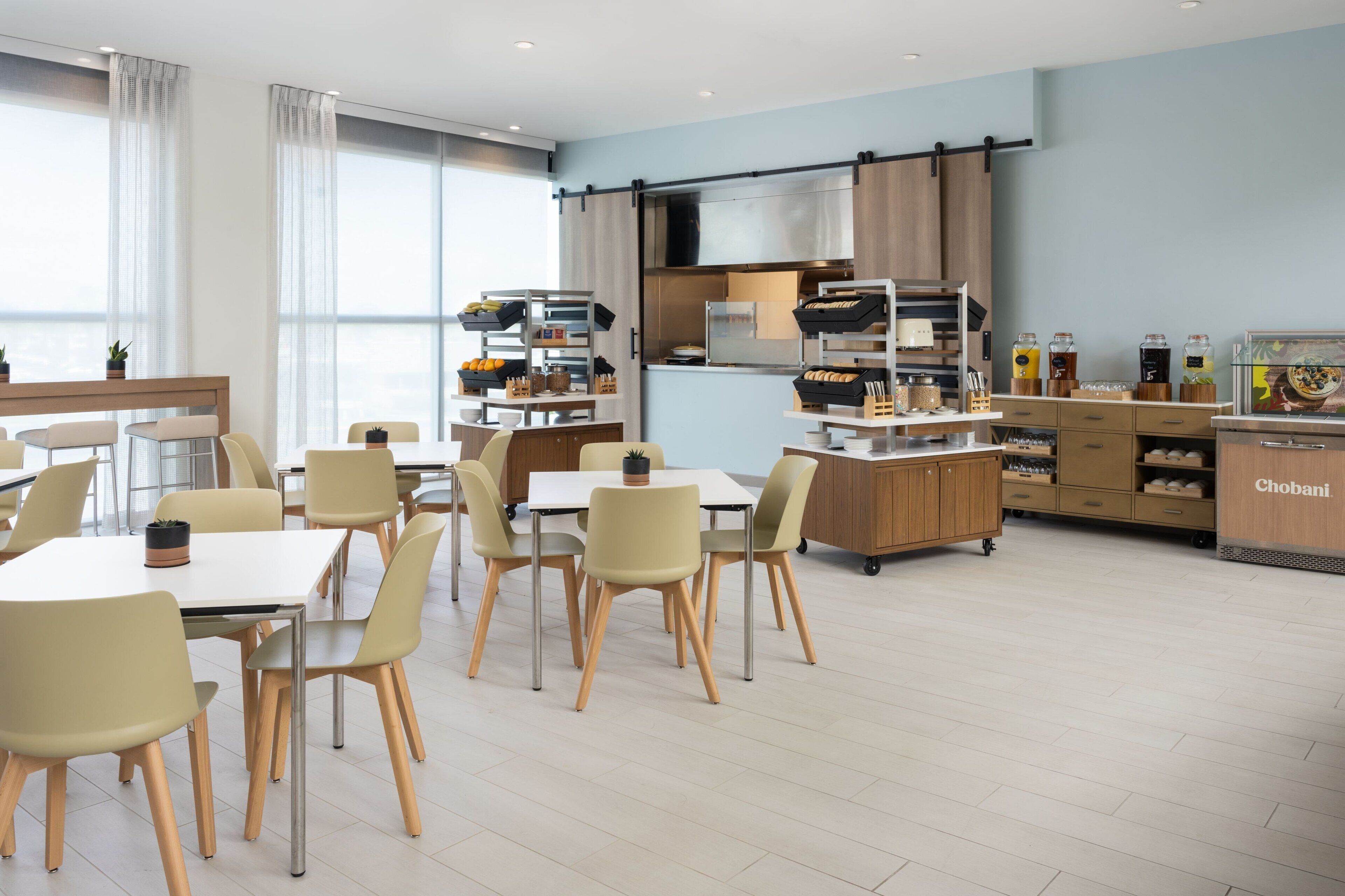 A modern dining area with tables and chairs, a kitchen area with appliances and storage, and a view through large windows.