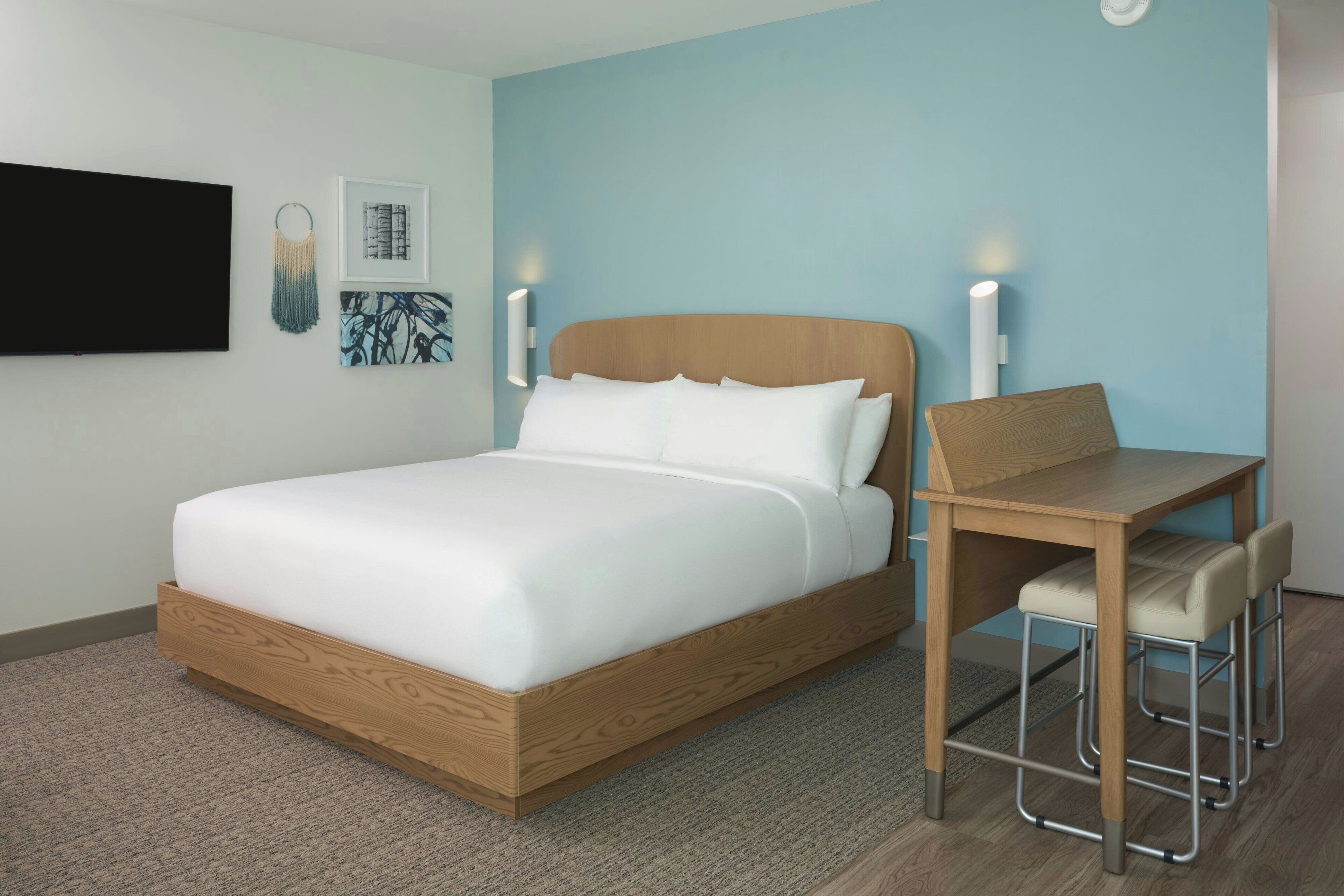 A modern hotel room with a wooden bed, a desk with two chairs, and a flat-screen TV mounted on the wall.