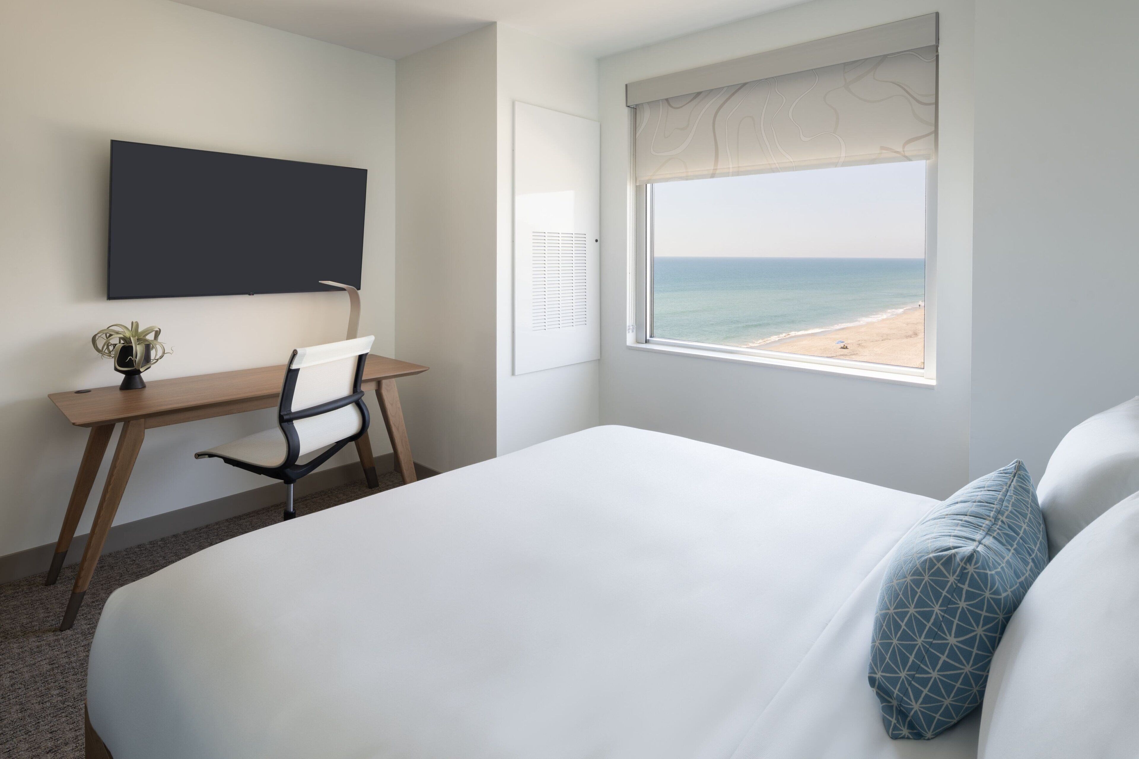 A hotel room with a bed, a desk, a chair, a TV, and a window with a beach view.