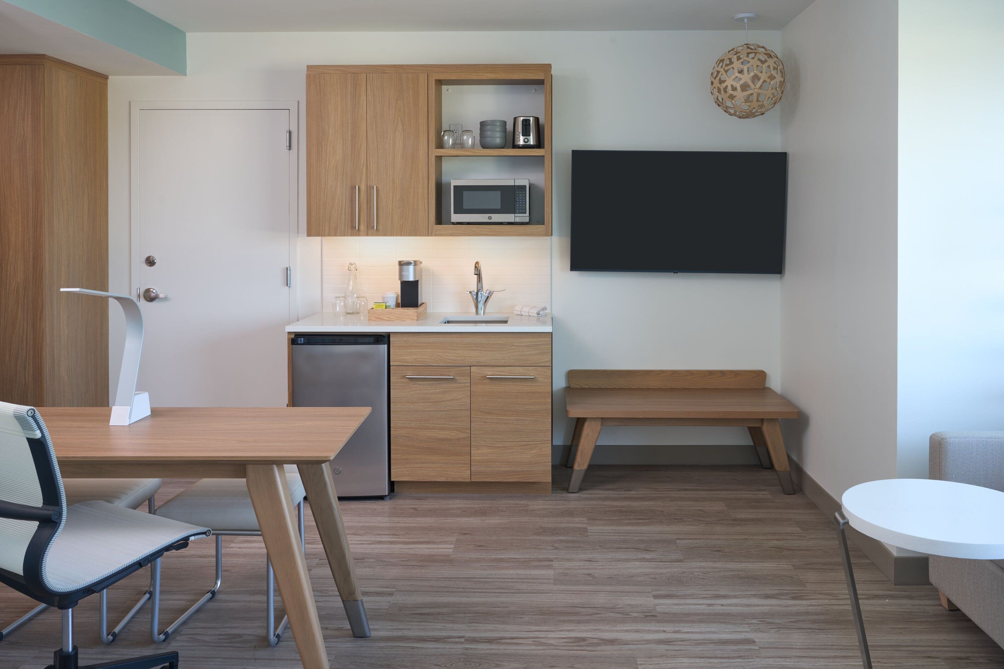 A modern kitchen with wooden cabinets, a microwave, and a sink. There is a dining table with chairs and a bench.