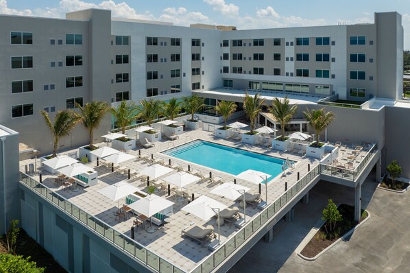 Área de piscina no terraço, com várias espreguiçadeiras e guarda-sóis, cercada por palmeiras e um edifício moderno com várias janelas.