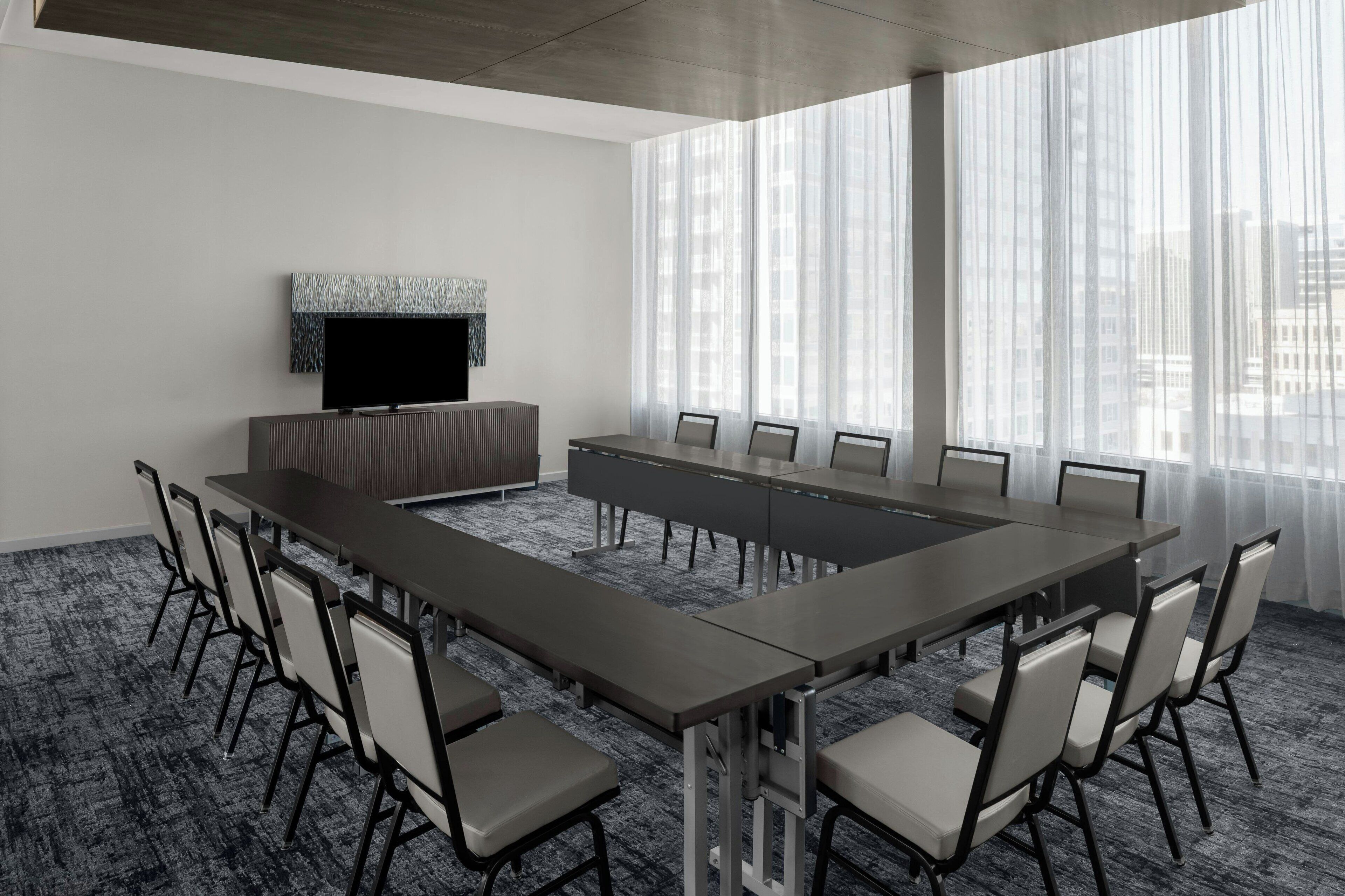 A conference room with a long table, chairs, a flat-screen TV, and large windows with sheer curtains.