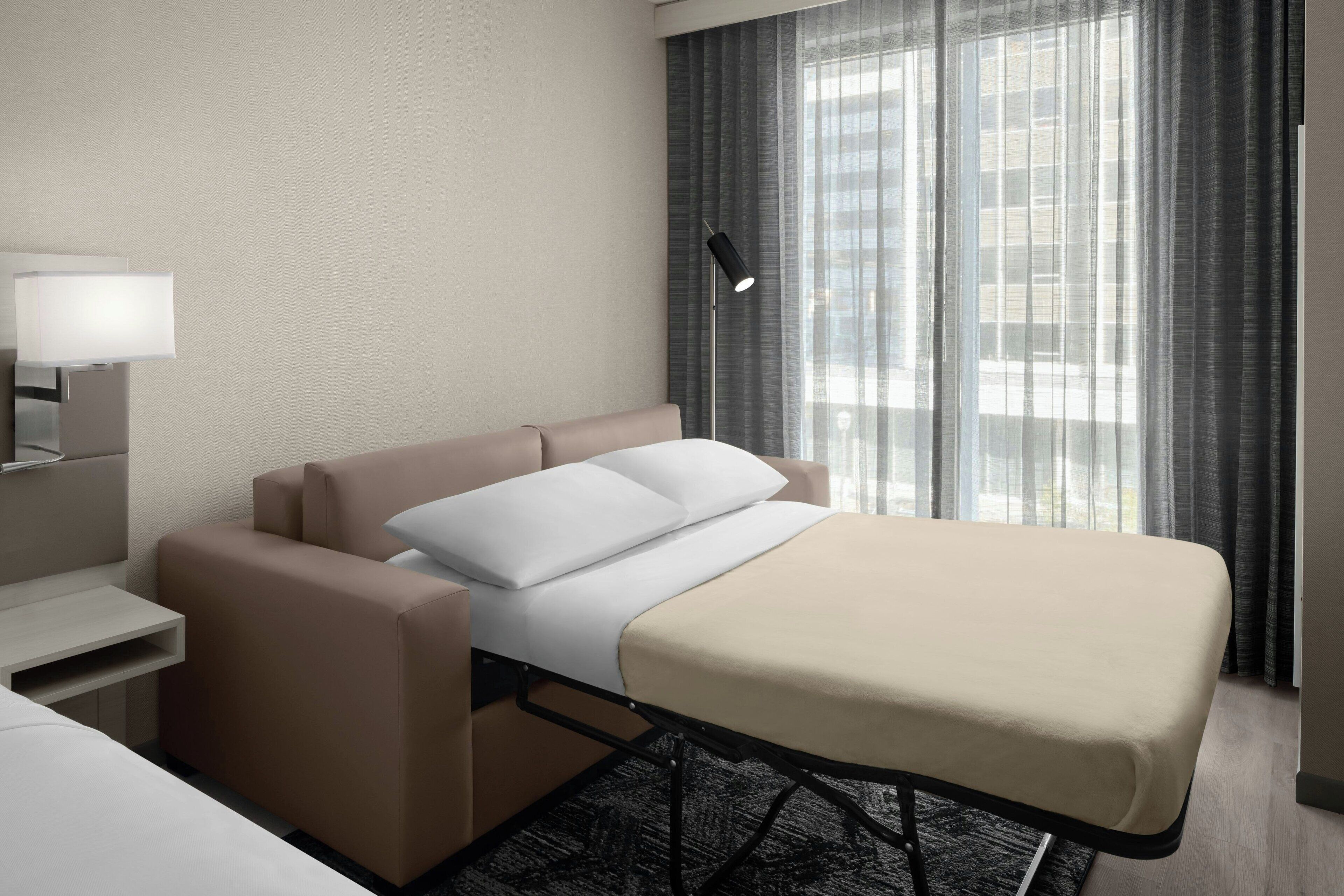 A modern hotel room with a bed, bedside table, lamp, and a view of the city through large windows.