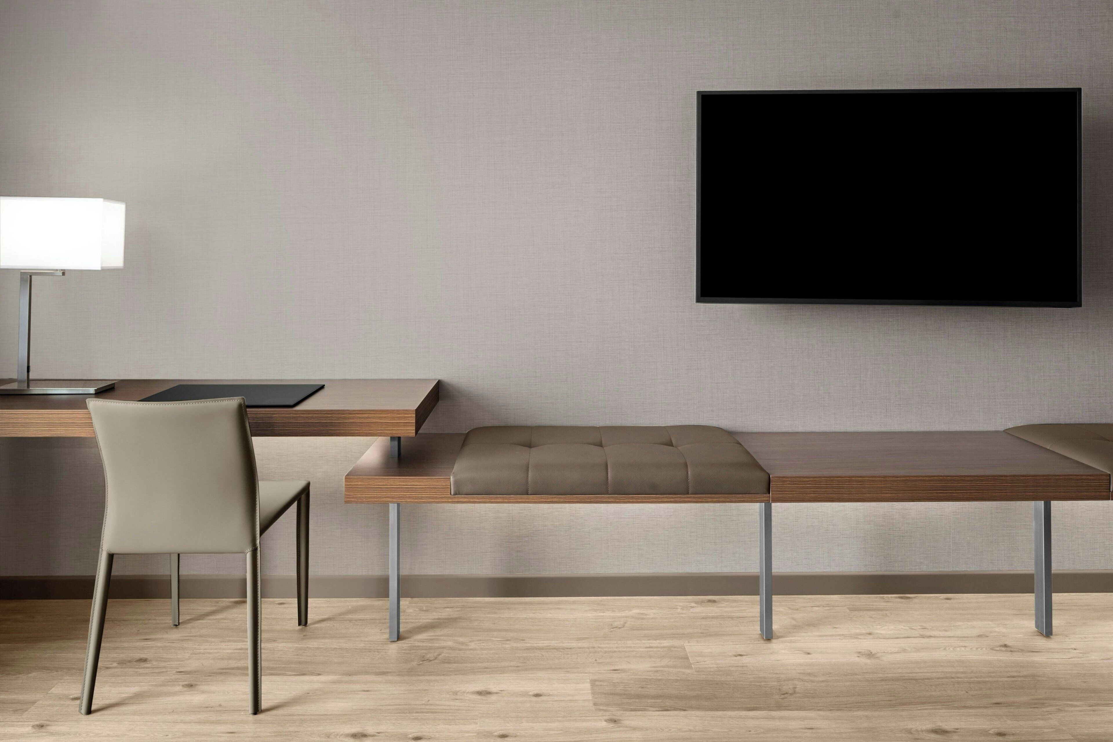 A modern room with a wooden desk, a cushioned bench, a chair, and a mounted flat-screen TV.