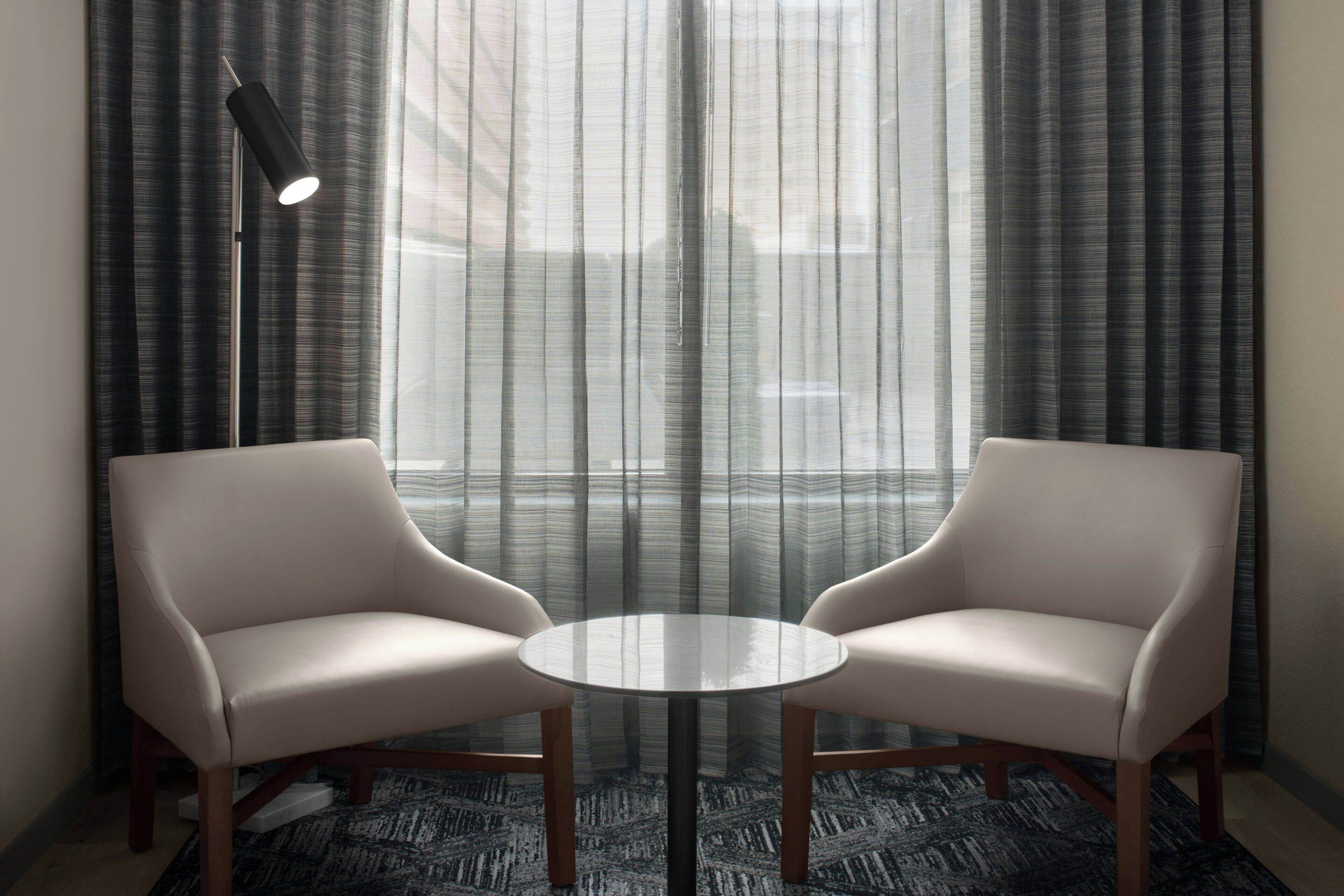 Quarto com duas poltronas de couro voltadas para uma mesa redonda de vidro, um abajur e vista através de cortinas leves.