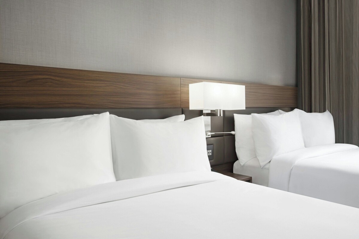 Een hotelkamer met een houten hoofdeinde, twee bedden met witte lakens, een nachtkastje en een lamp.