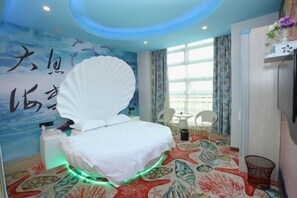 Hotelkamer met een bed, een grote schelpdecoratie, een muurschildering met zeedieren, een raam met gordijnen, en een stoel.