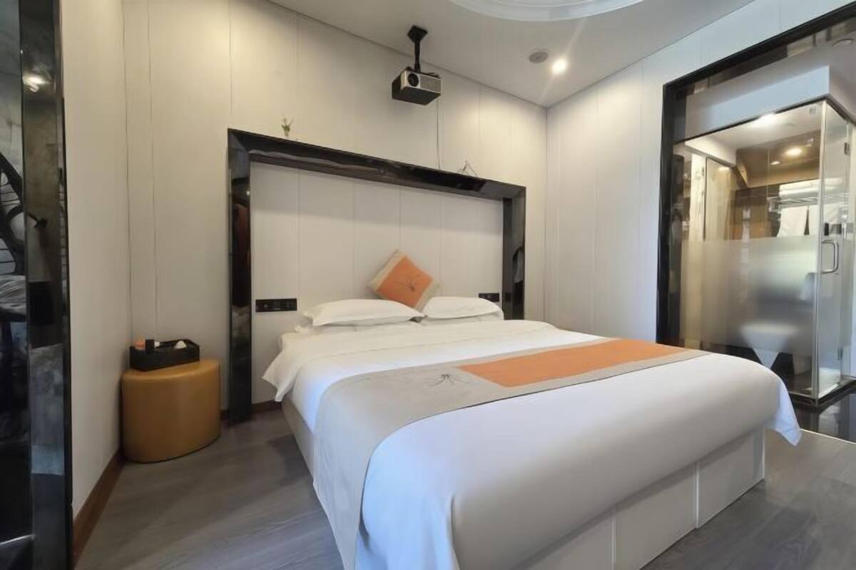 Une chambre d’hôtel moderne dotée d’un grand lit, d’un petit pouf rond, d’un projecteur au plafond et d’une douche en verre.