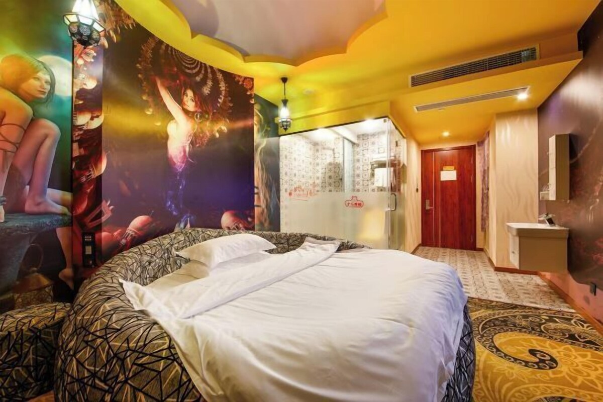 Une chambre d’hôtel avec un grand lit, une salle de bain avec douche et toilettes, et un mur orné d’une affiche.
