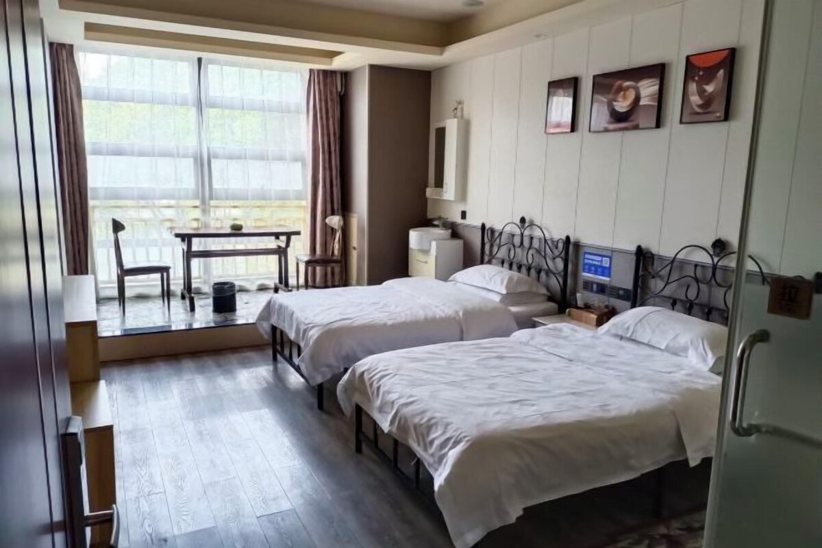 Une chambre d’hôtel avec deux lits, un bureau et une chaise. Il y a une fenêtre avec des rideaux, un téléviseur fixé au mur et des tableaux encadrés.