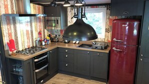 A compact kitchen with a gas stove, oven, and a red refrigerator. There is a wooden floor and a window with a view of greenery. - Pet Friendly Lodge - Sleeps 6 (Fordingbridge)