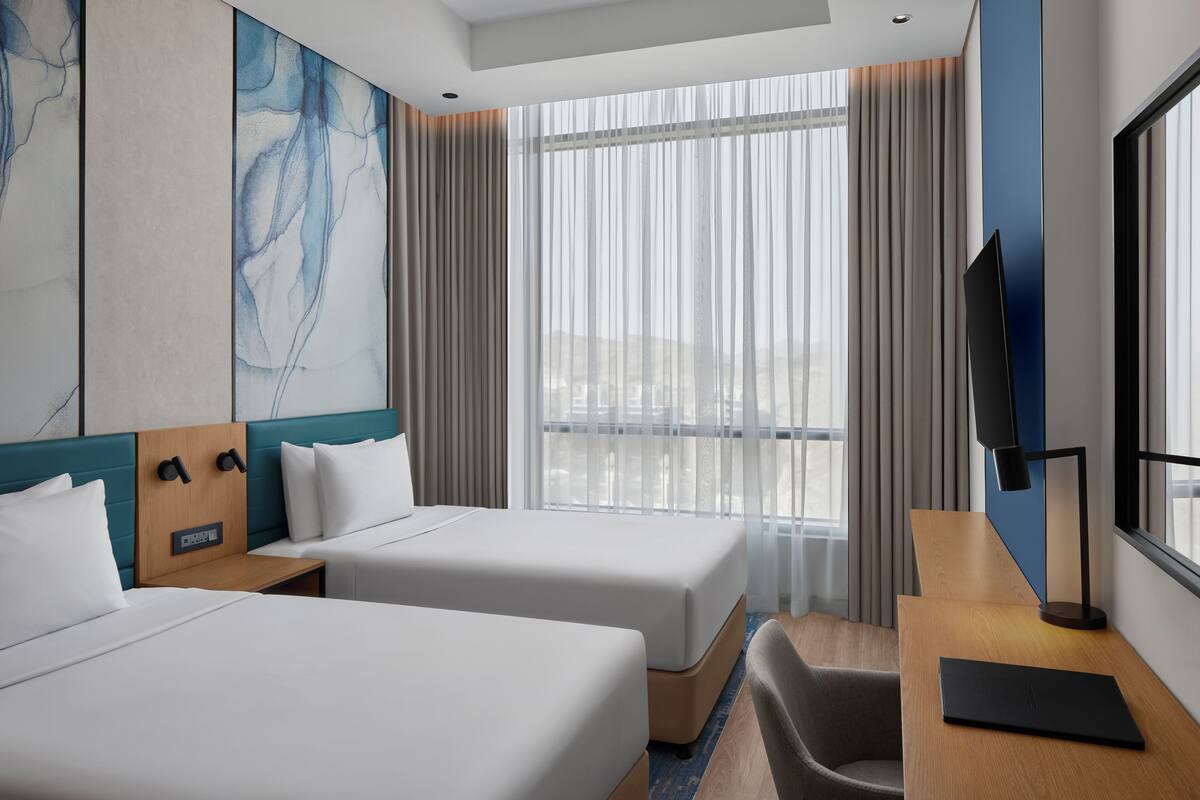 Habitación de hotel con dos camas, un escritorio con una laptop, un televisor y un amplio ventanal con cortinas.