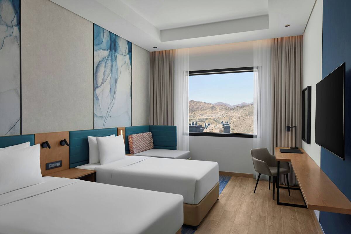 Habitación de hotel con dos camas, un escritorio, una silla, un televisor y un ventanal con vistas a la montaña.