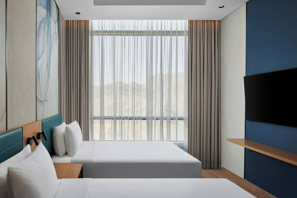 Habitación de hotel con dos camas, un ventanal con cortinas, un televisor montado y un cuadro en la pared.