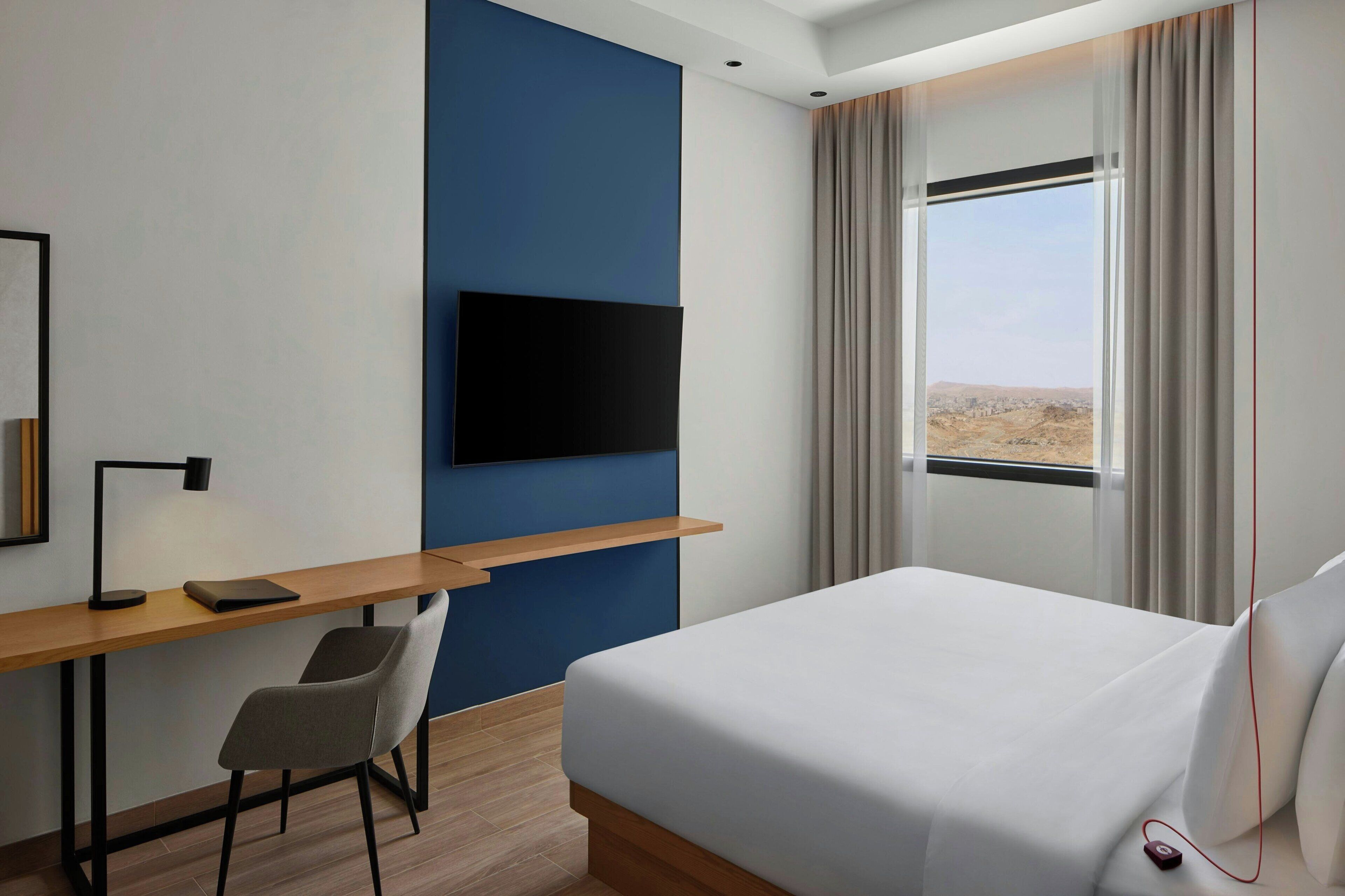A hotel room with a bed, a desk with a chair, a flat-screen TV mounted on the wall, and a window with curtains.