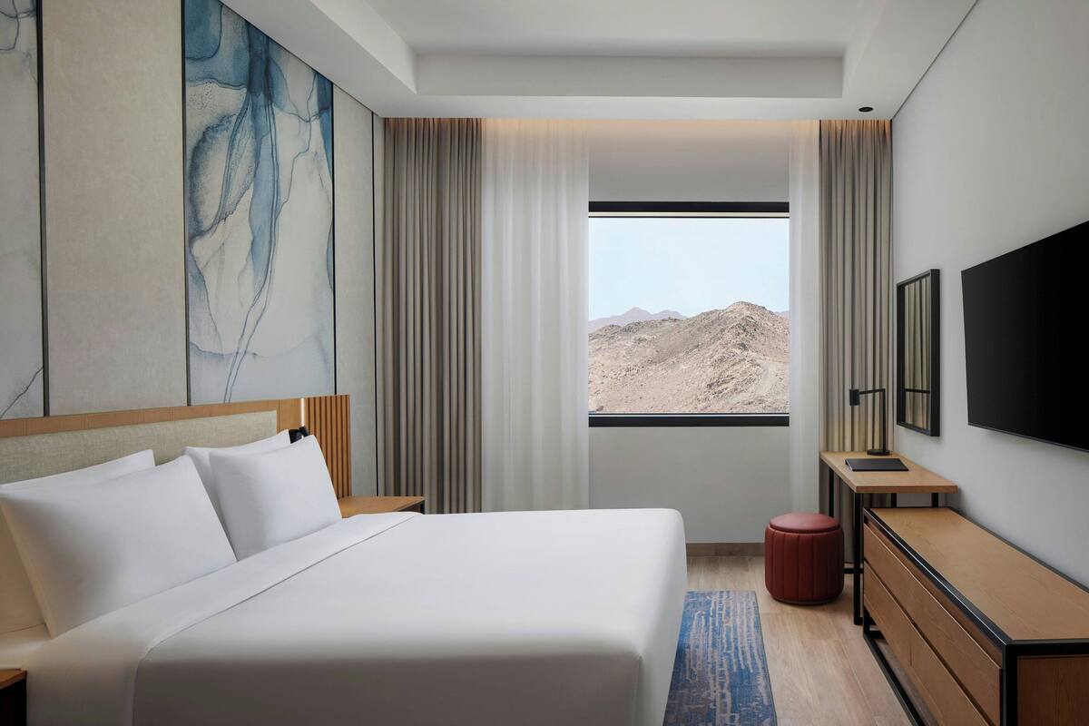 Habitación de hotel con una cama grande, un escritorio, un televisor y una ventana con vistas a un paisaje montañoso.