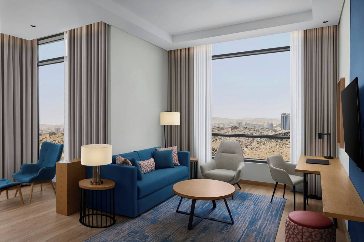 Una habitación de hotel moderna con un sofá, sillas, una mesa de centro, un televisor y ventanas amplias que ofrecen vistas de la ciudad.