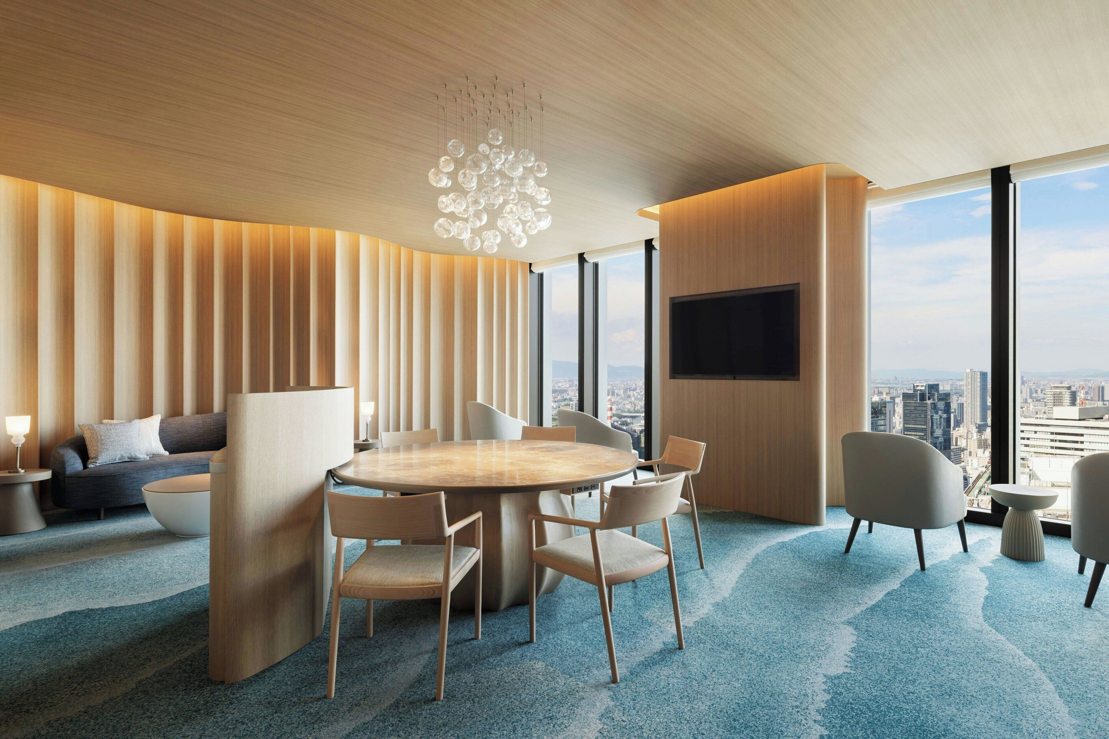 A modern hotel room with a large round table, comfortable seating, a flat-screen TV, and a city view.