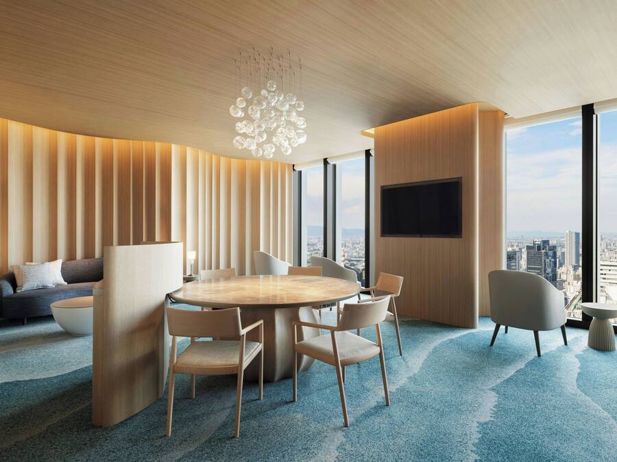 A modern hotel room with a large round table, comfortable seating, a flat-screen TV, and a city view.