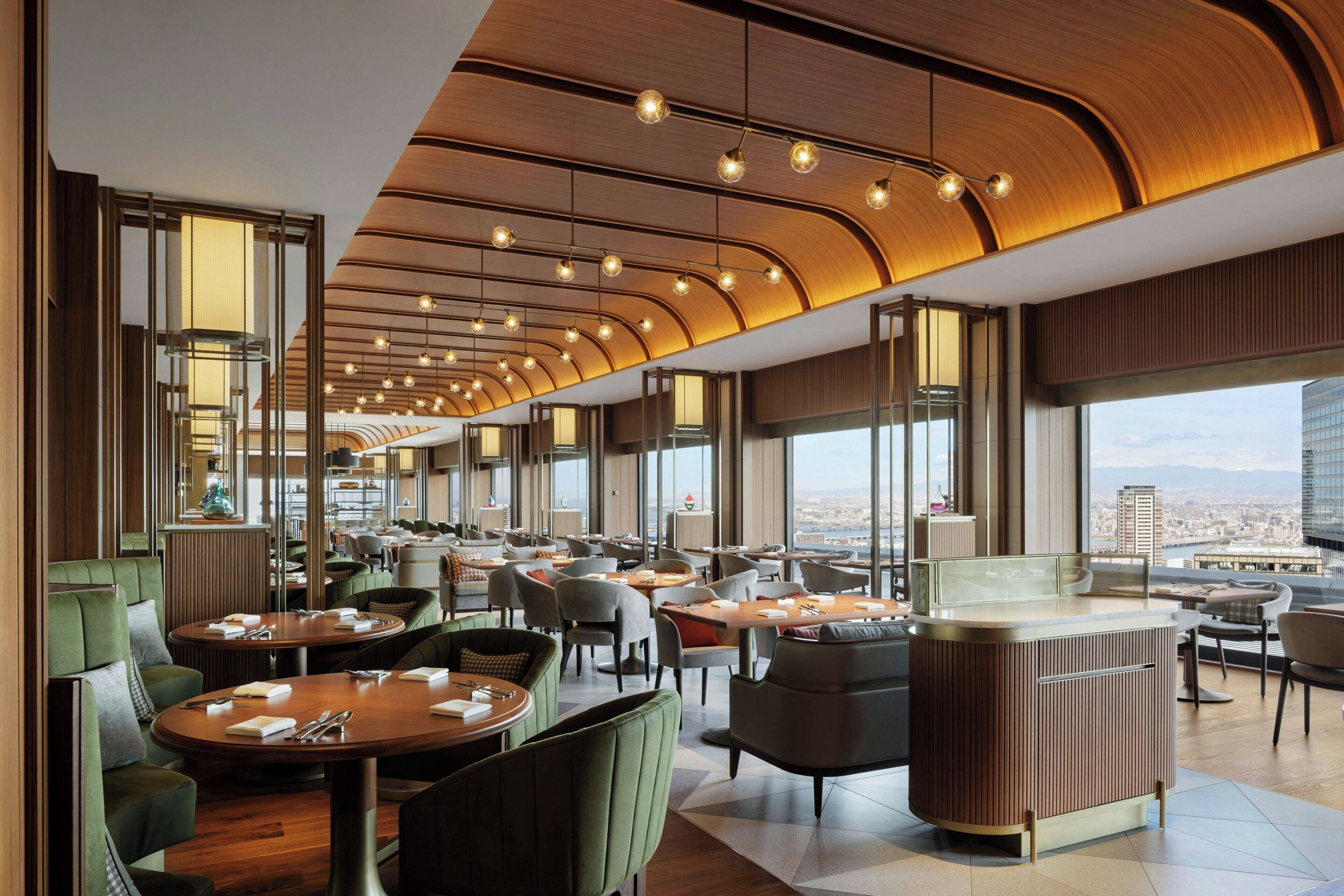 Un restaurant moderne avec des tables rondes, des sièges rembourrés de tissu vert et une vue sur le paysage urbain grâce à de grandes fenêtres.
