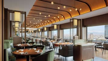 Un restaurant moderne avec des tables rondes, des sièges rembourrés de tissu vert et une vue sur le paysage urbain grâce à de grandes fenêtres.