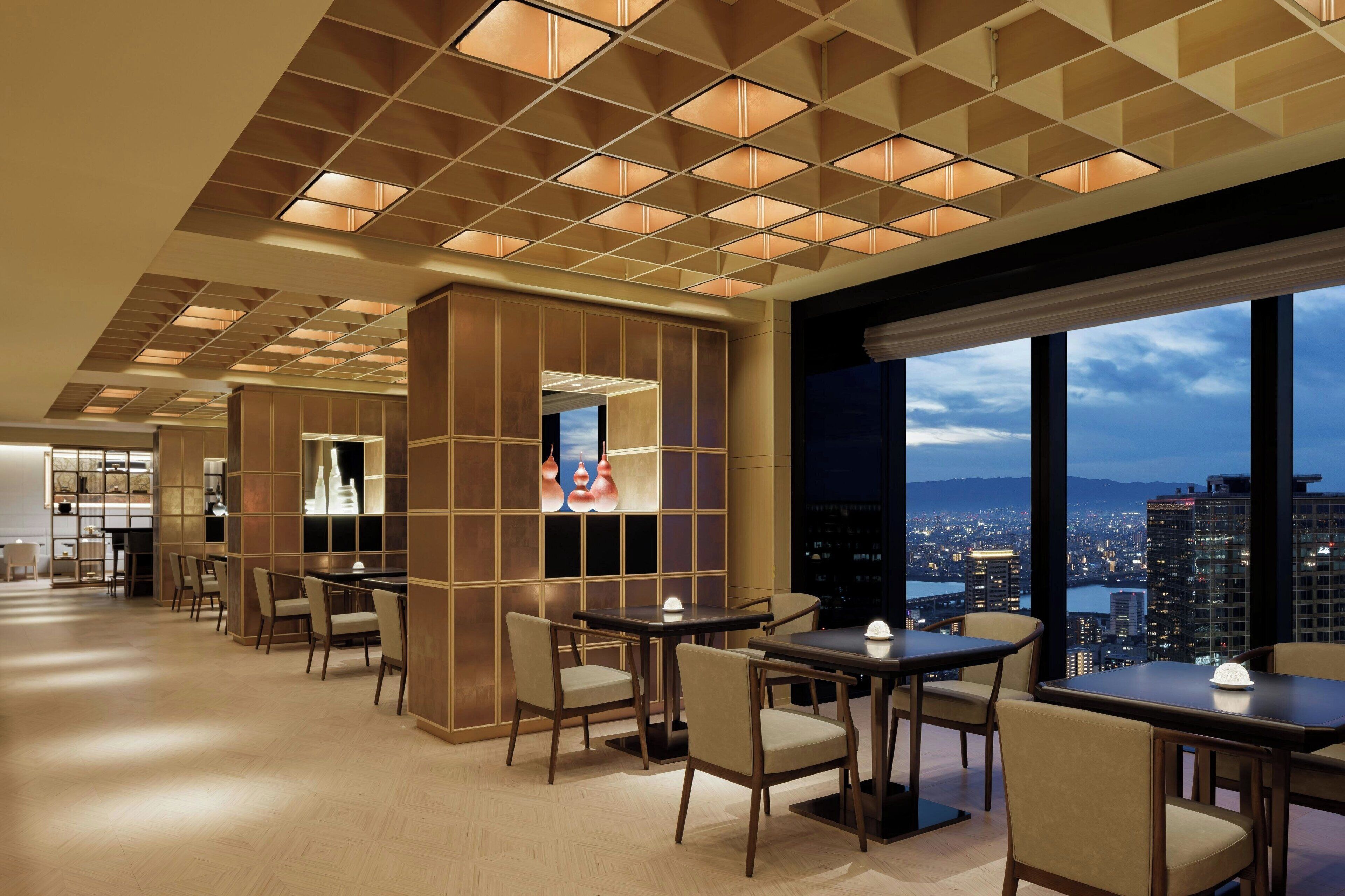 A modern hotel lobby with a city view, featuring a spacious seating area with tables and chairs, a decorative wall with a television, and a ceiling with recessed lighting.