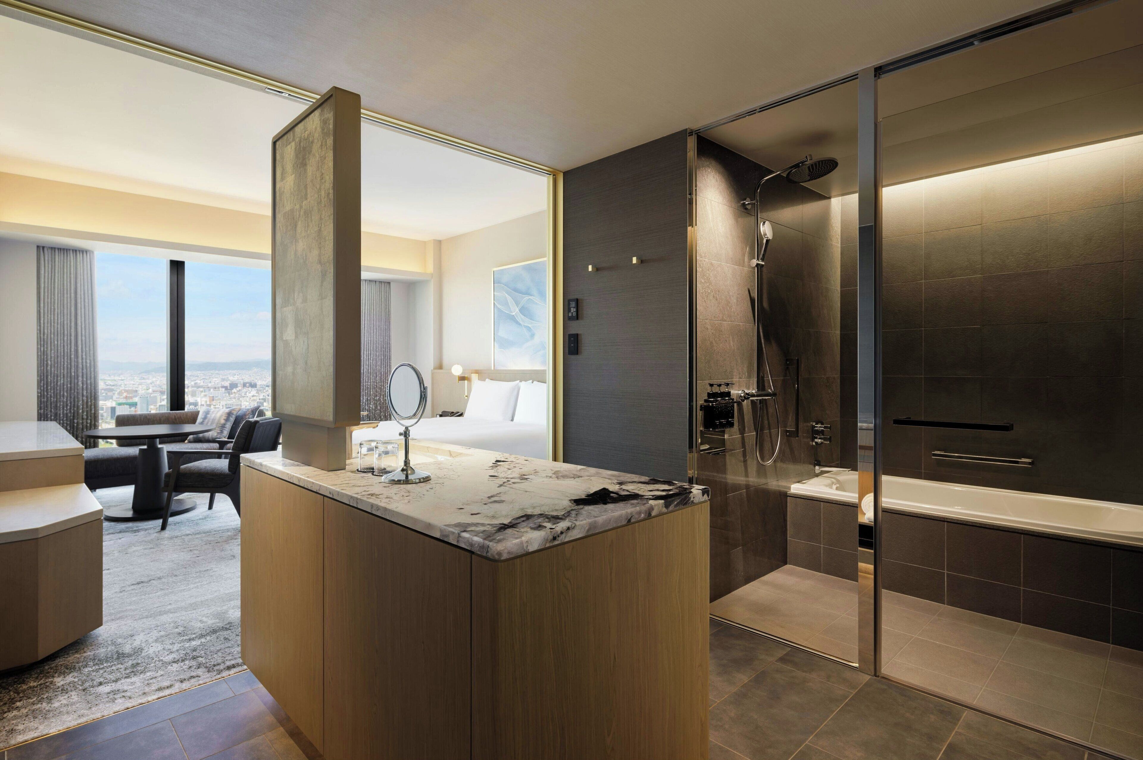 A modern hotel room with a large marble island, a separate shower area, a sitting area with a view, and a large window.