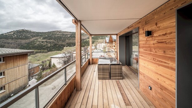 A balcony with wooden flooring, a glass railing, and a view of a hilly landscape.