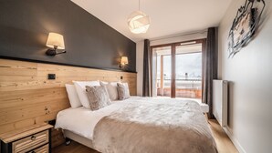 A modern bedroom with a wooden headboard, a large bed with a beige blanket, two wall-mounted lamps, and a view of a cityscape through a large window.