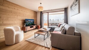 A modern living room with a wooden wall, a large window with a city view, a sofa, a chair, a coffee table, and a TV.
