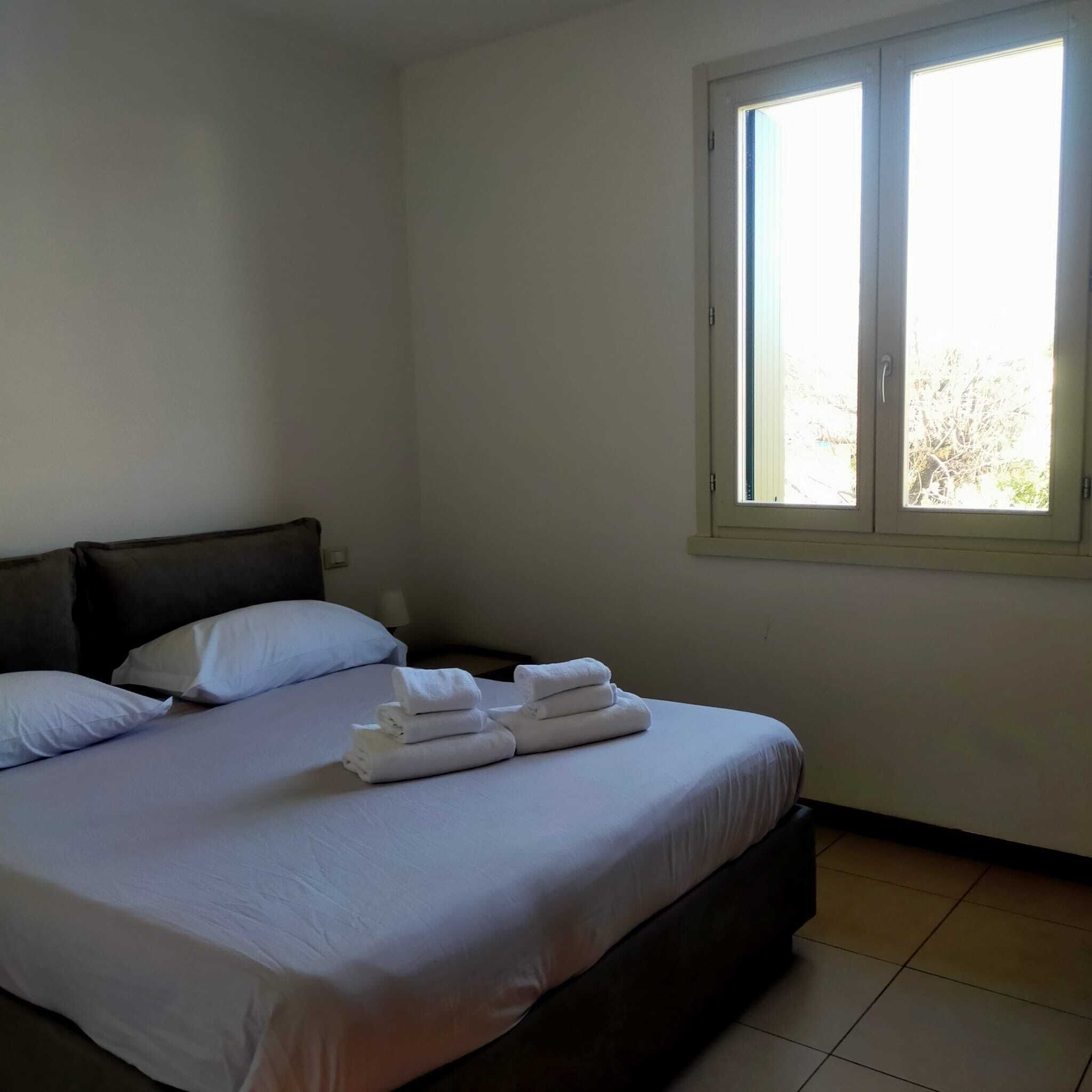 A neatly made bed with white linens and towels, a dark headboard, and a window with blinds.