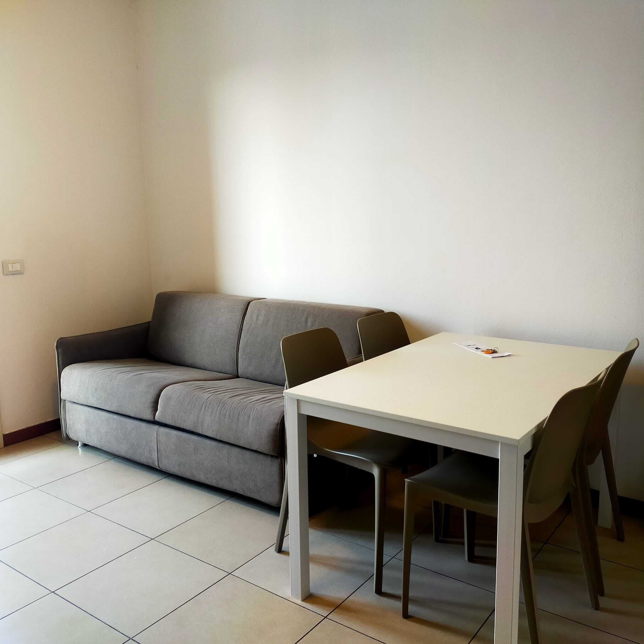 Ein kleiner, moderner Raum mit einem grauen Sofa, einem weißen Tisch und zwei Stühlen.
