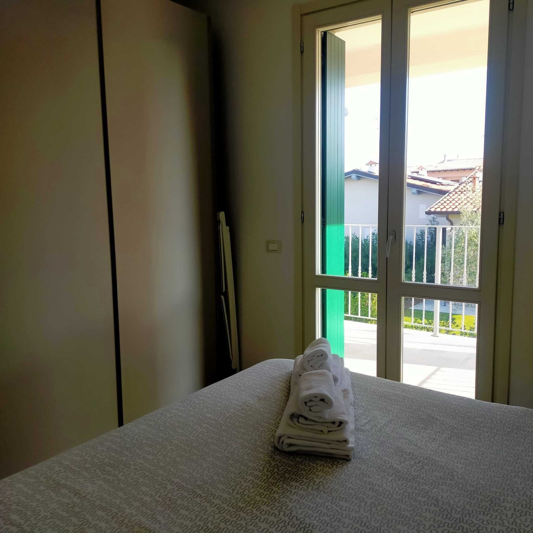 Habitación de hotel con cama, toalla doblada, armario y puerta corrediza de vidrio que da a un espacio exterior con vistas a edificios y vegetación.