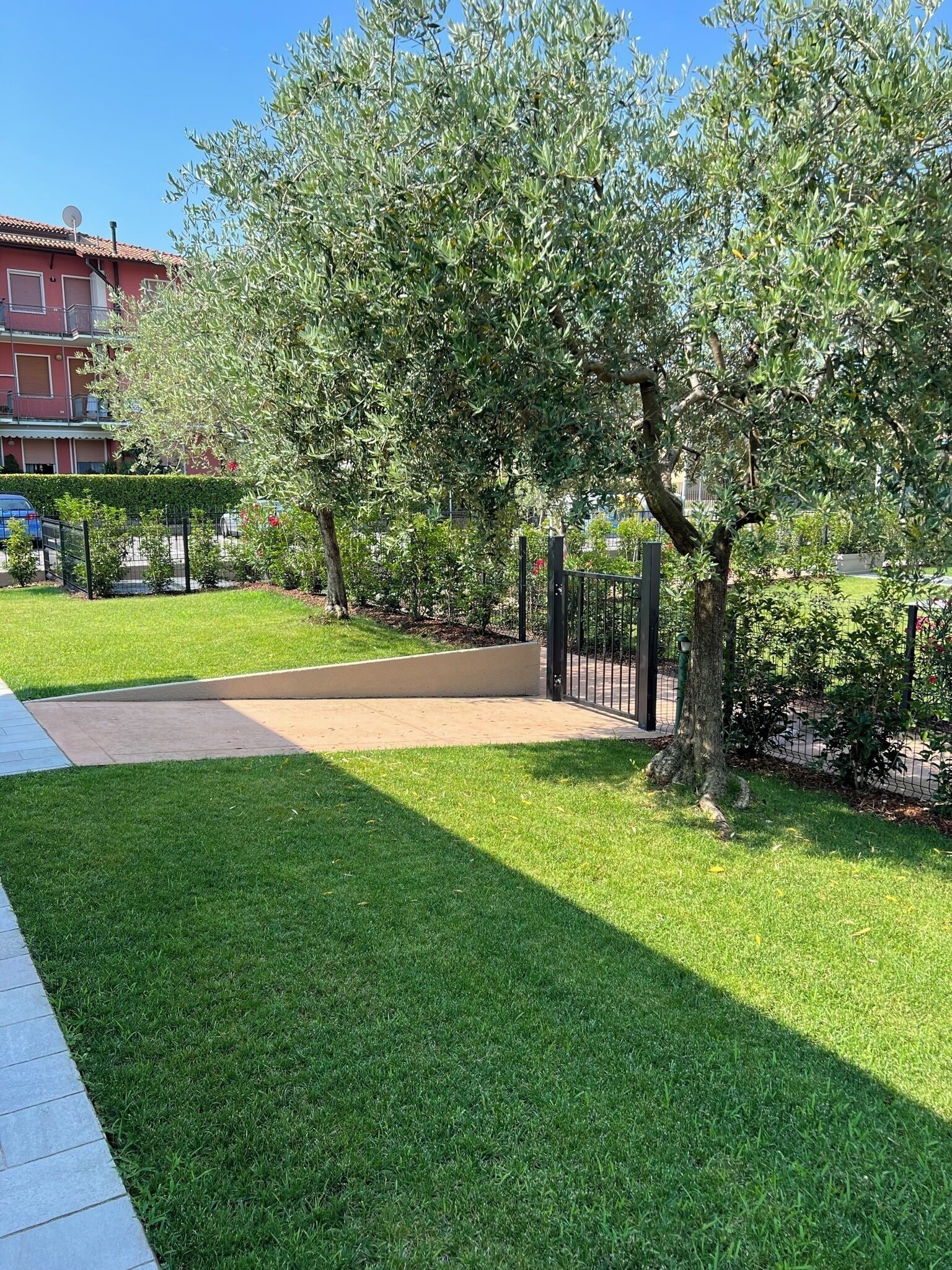 Una zona verda ben cuidada amb un camí pavimentat, una tanca i un edifici al fons.