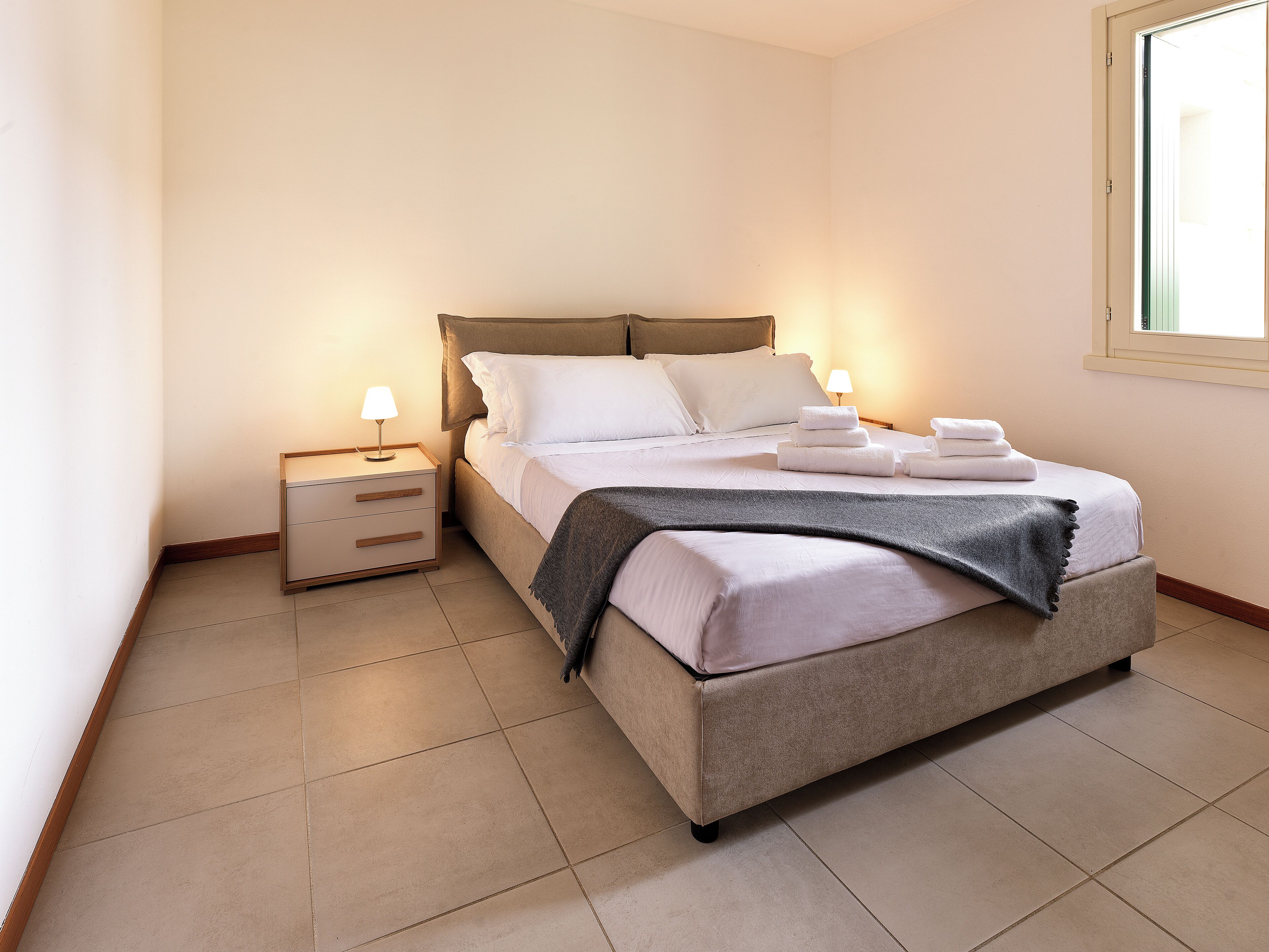 A neatly made bed with white linens and a grey throw blanket, a bedside table with a lamp, and a window with blinds.