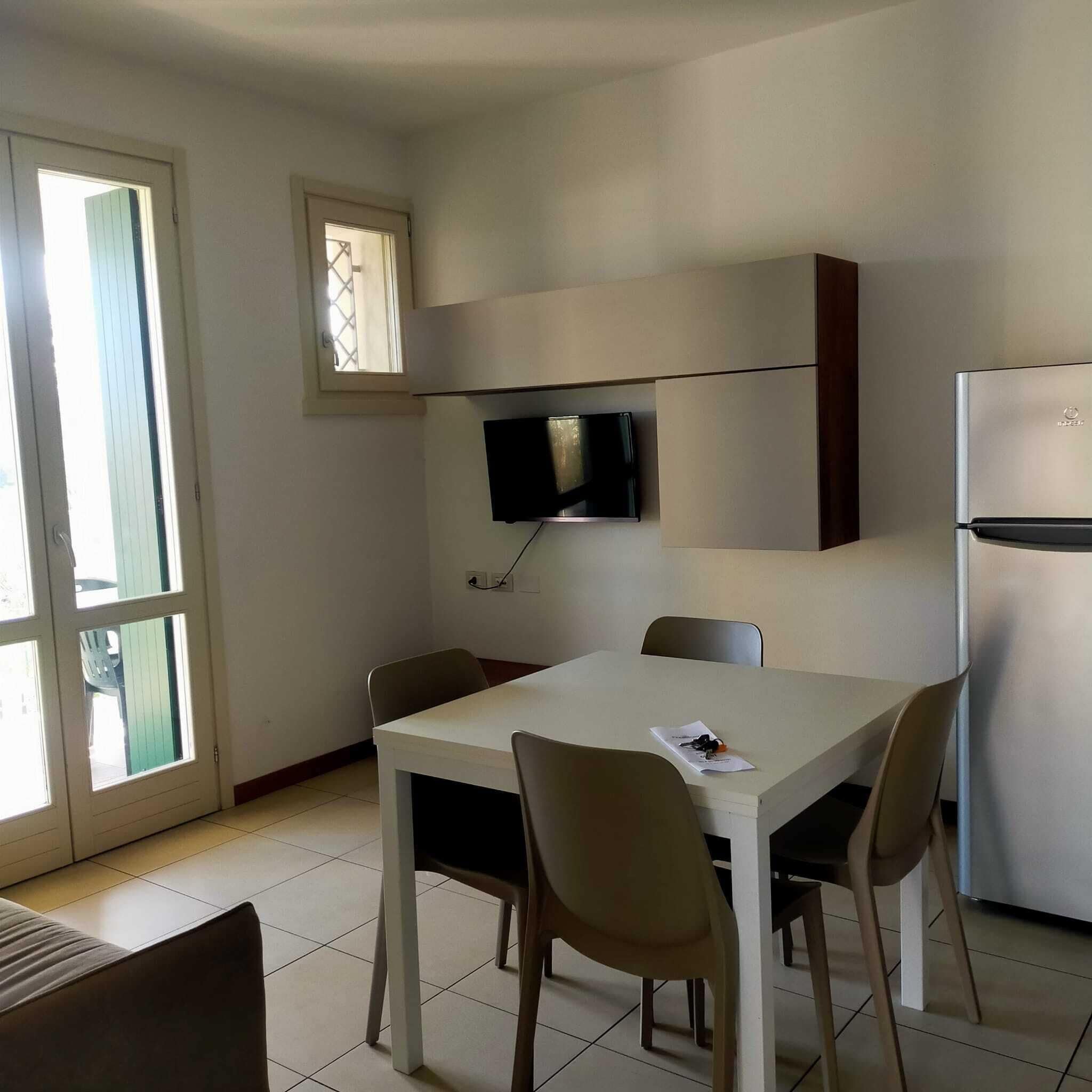 Una cocina pequeña y moderna con una mesa y sillas blancas, televisor integrado y refrigerador.