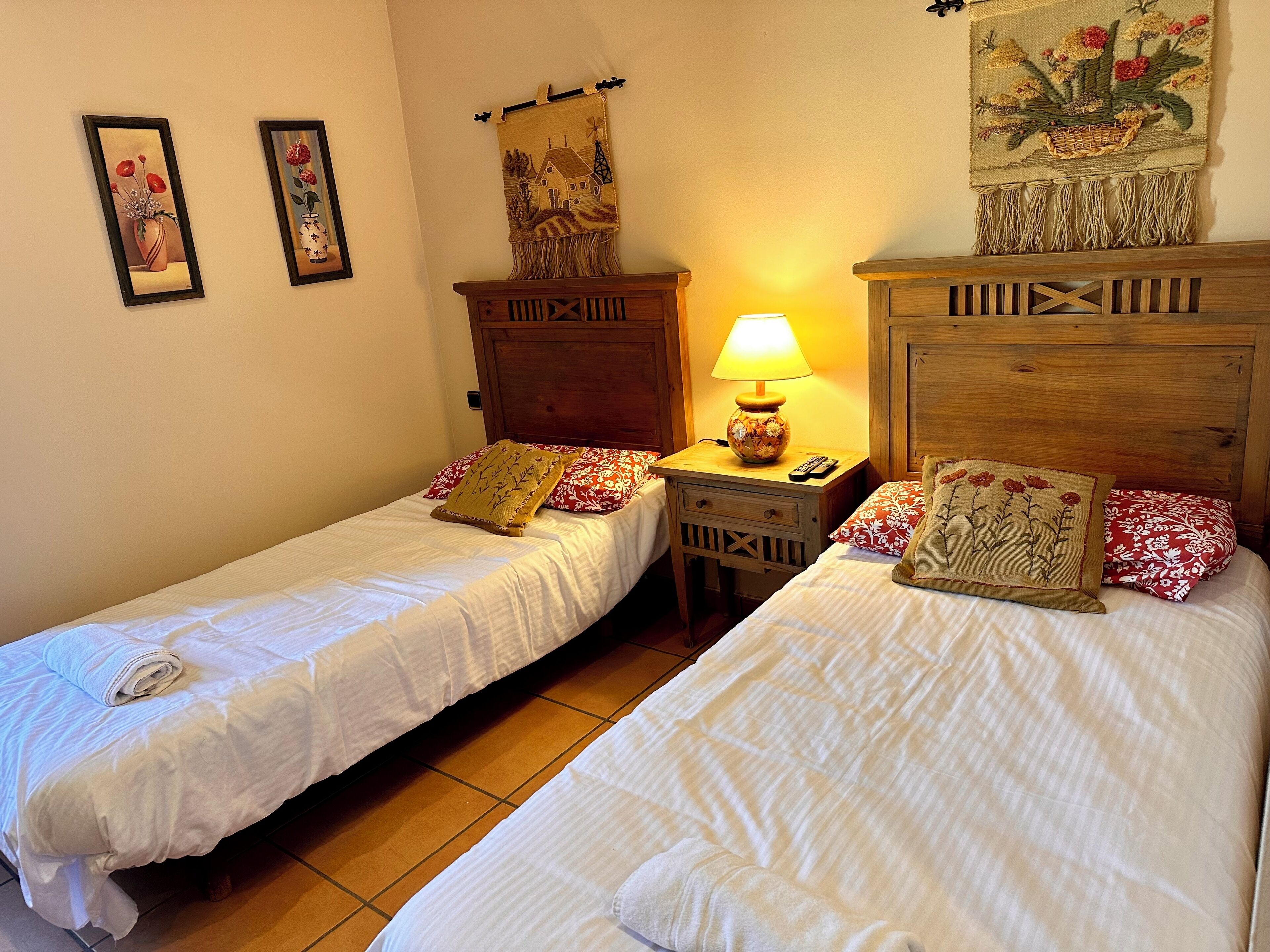 A room with two single beds, a wooden nightstand with a lamp, and a wall-mounted tapestry.