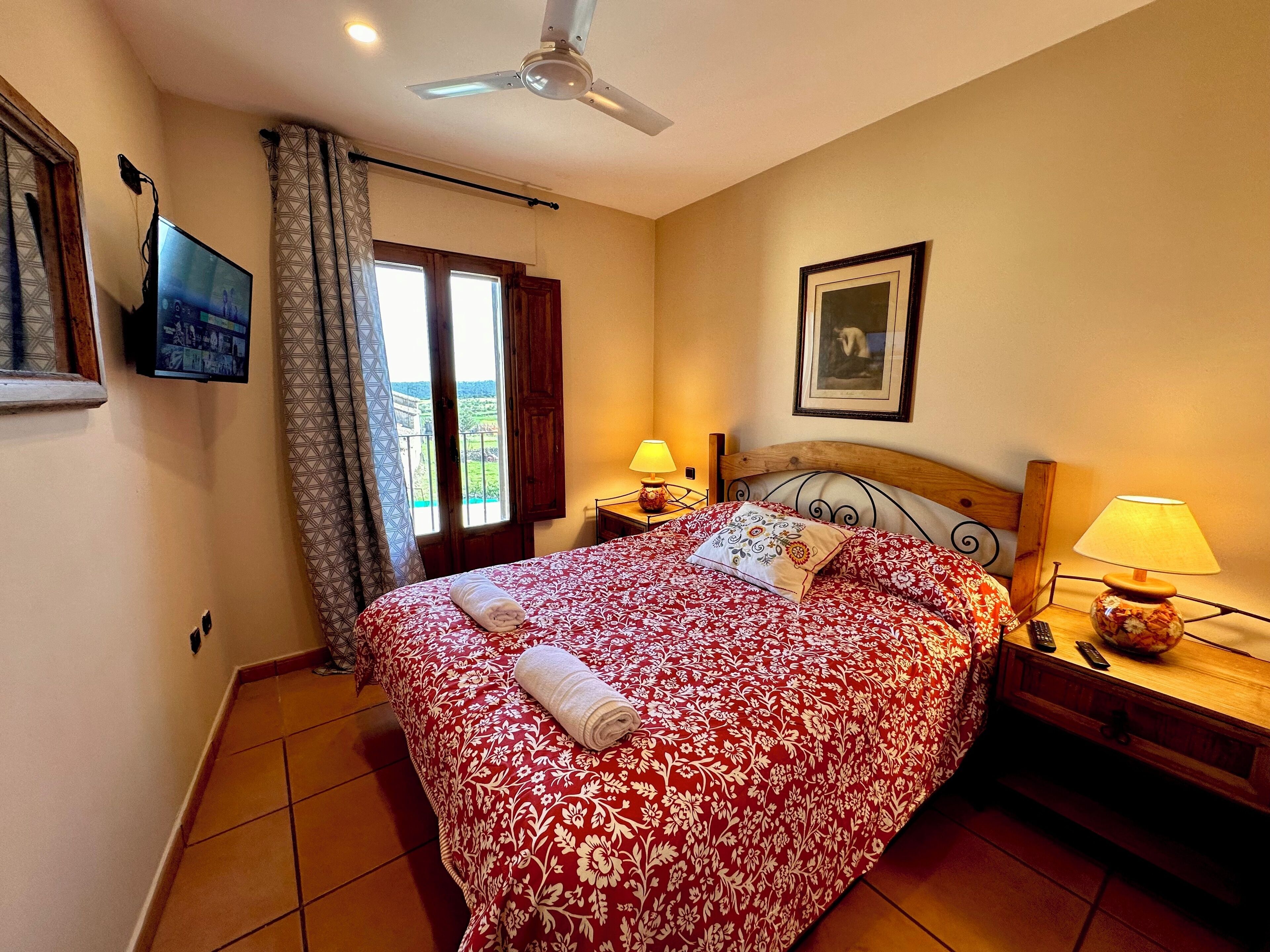 Un dormitorio con cama, dos mesitas de noche con lámparas, ventilador de techo, televisor montado en la pared, ventana con cortinas e imagen enmarcada en la pared.