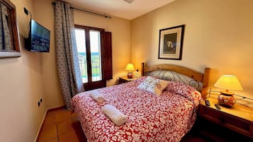 A bedroom with a bed, two bedside tables with lamps, a ceiling fan, a TV mounted on the wall, a window with curtains, and a framed picture on the wall.